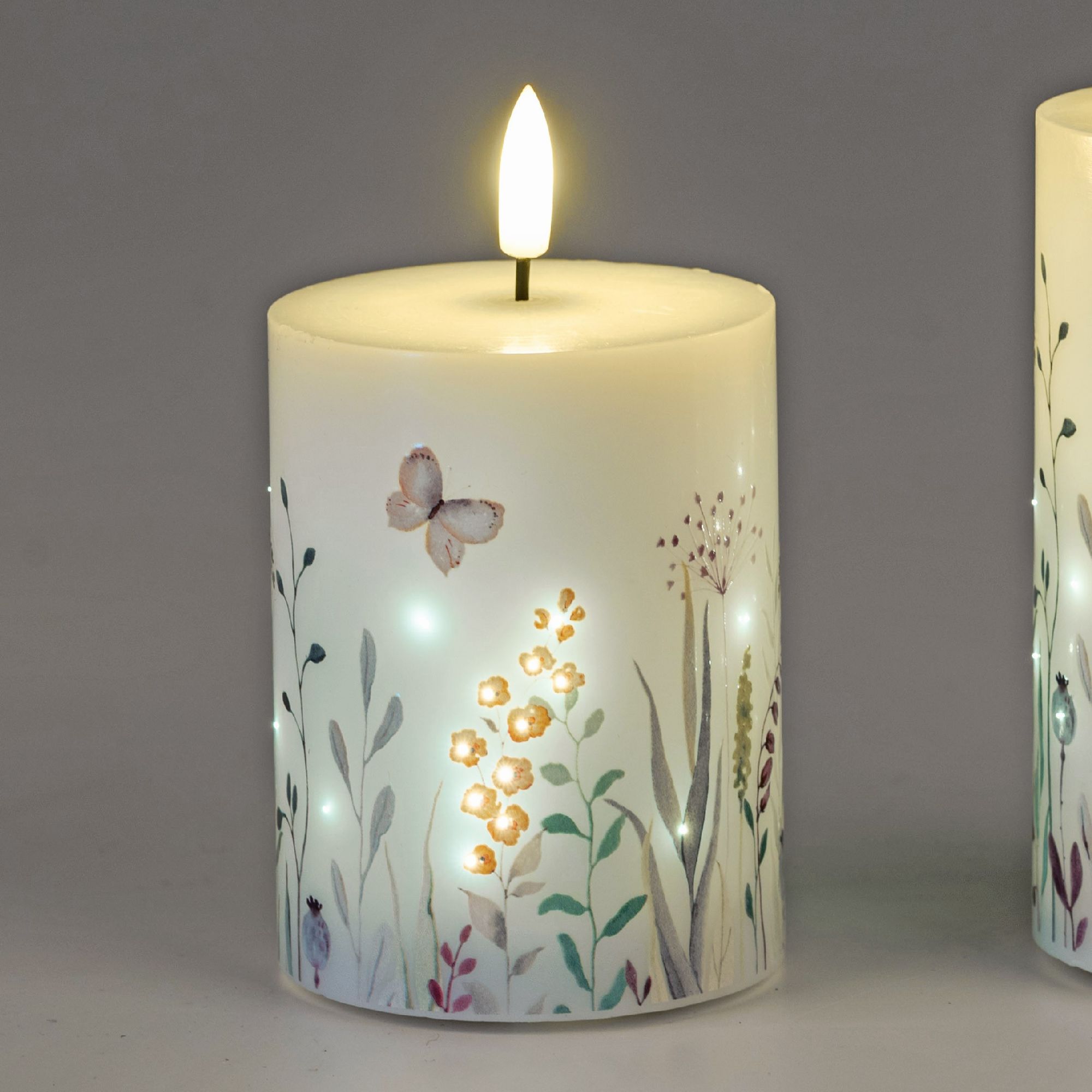 Eine weiße Kerze mit floralen Motiven und einer flackernden Flamme. Im Hintergrund sind weitere Kerz