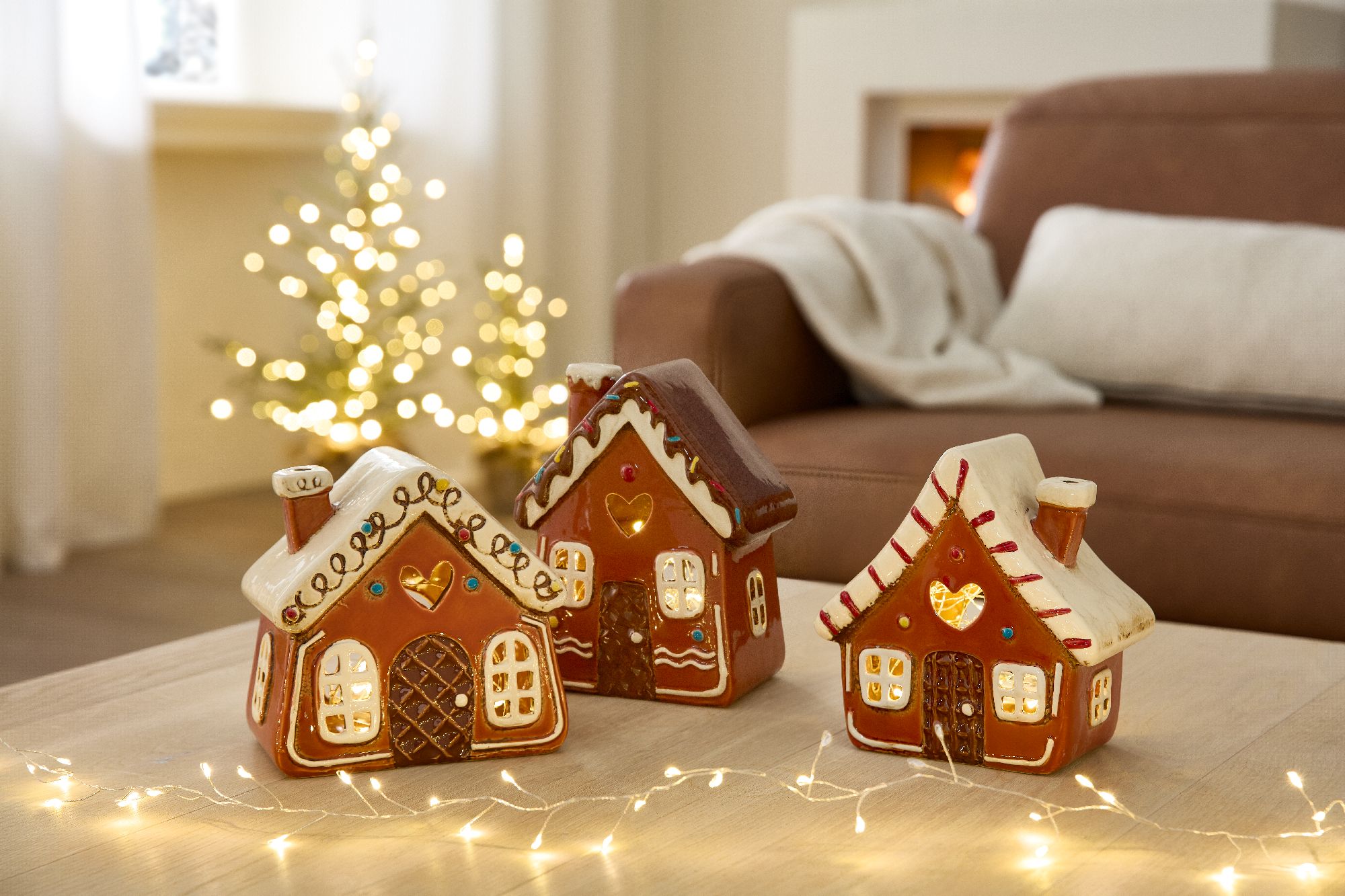 Drei dekorative Lebkuchenhäuser stehen auf einem Tisch, im Hintergrund ein Weihnachtsbaum mit Lichte