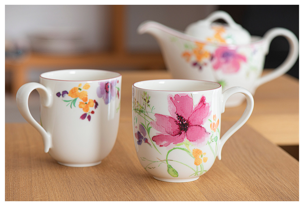 Zwei Tassen mit bunten Blumenmustern und eine Teekanne auf einem Holztisch. Hintergrund unscharf.