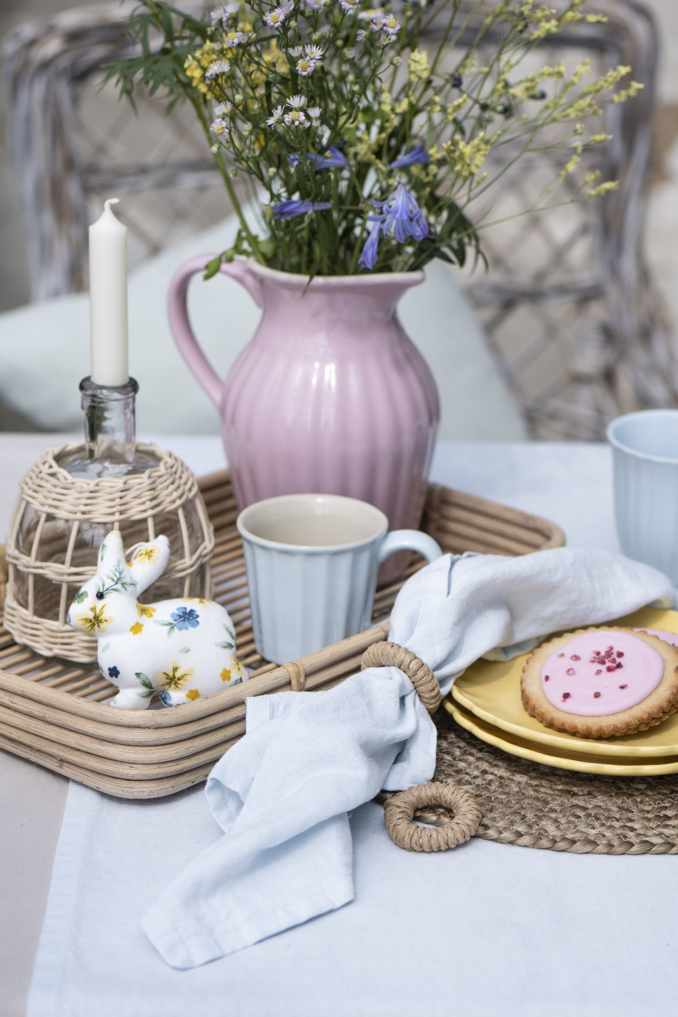 Ein Tischgedeck mit einer rosa Kanne, Blumen, Geschirr, einem Hasen aus Keramik, Serviette und einem Keks auf einem Tablett.