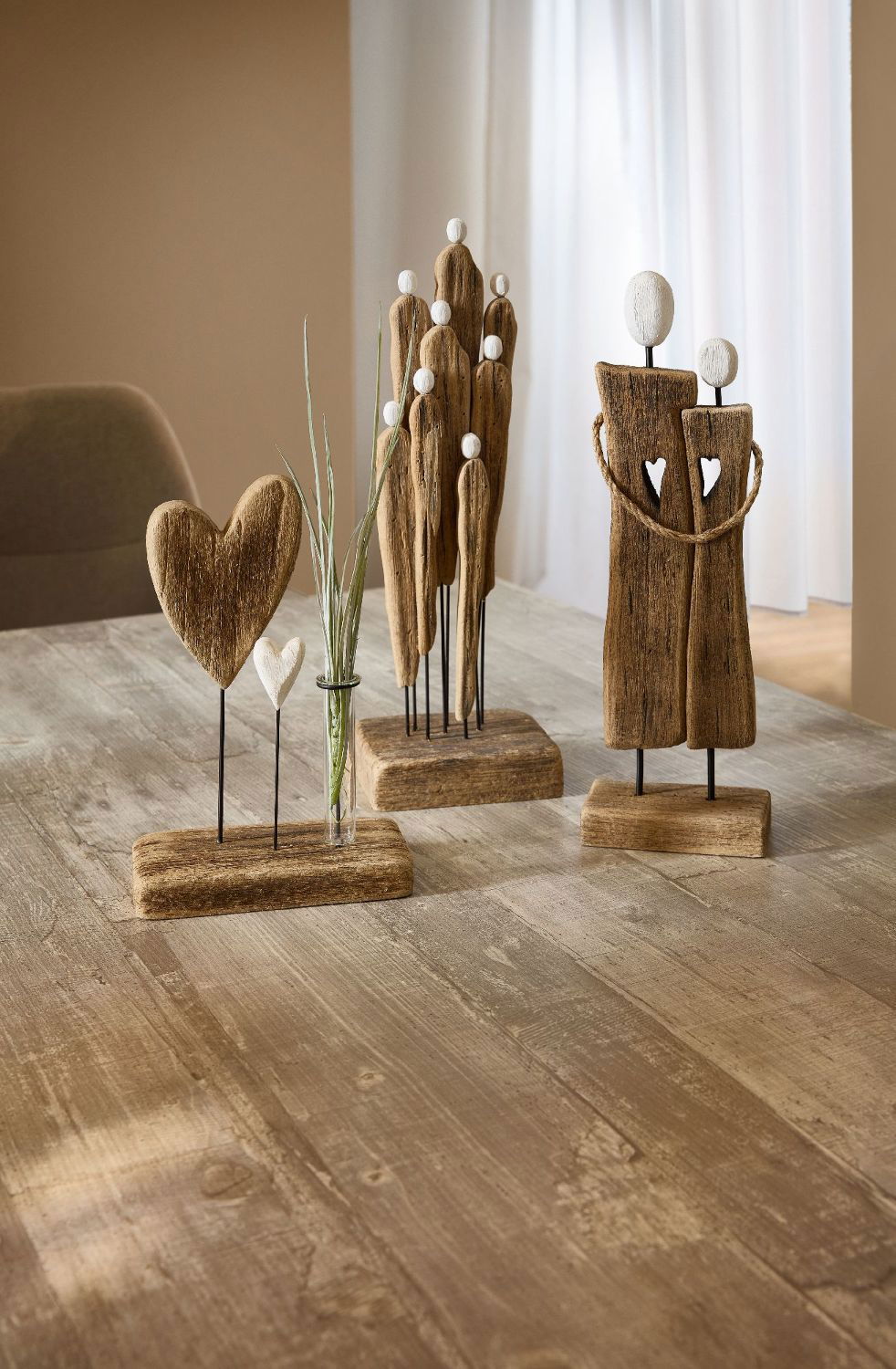 Drei dekorative Figuren aus Holz stehen auf einem Tisch: ein Herz, eine Gruppe von Menschen und ein Paar, umgeben von einer Pflanze.