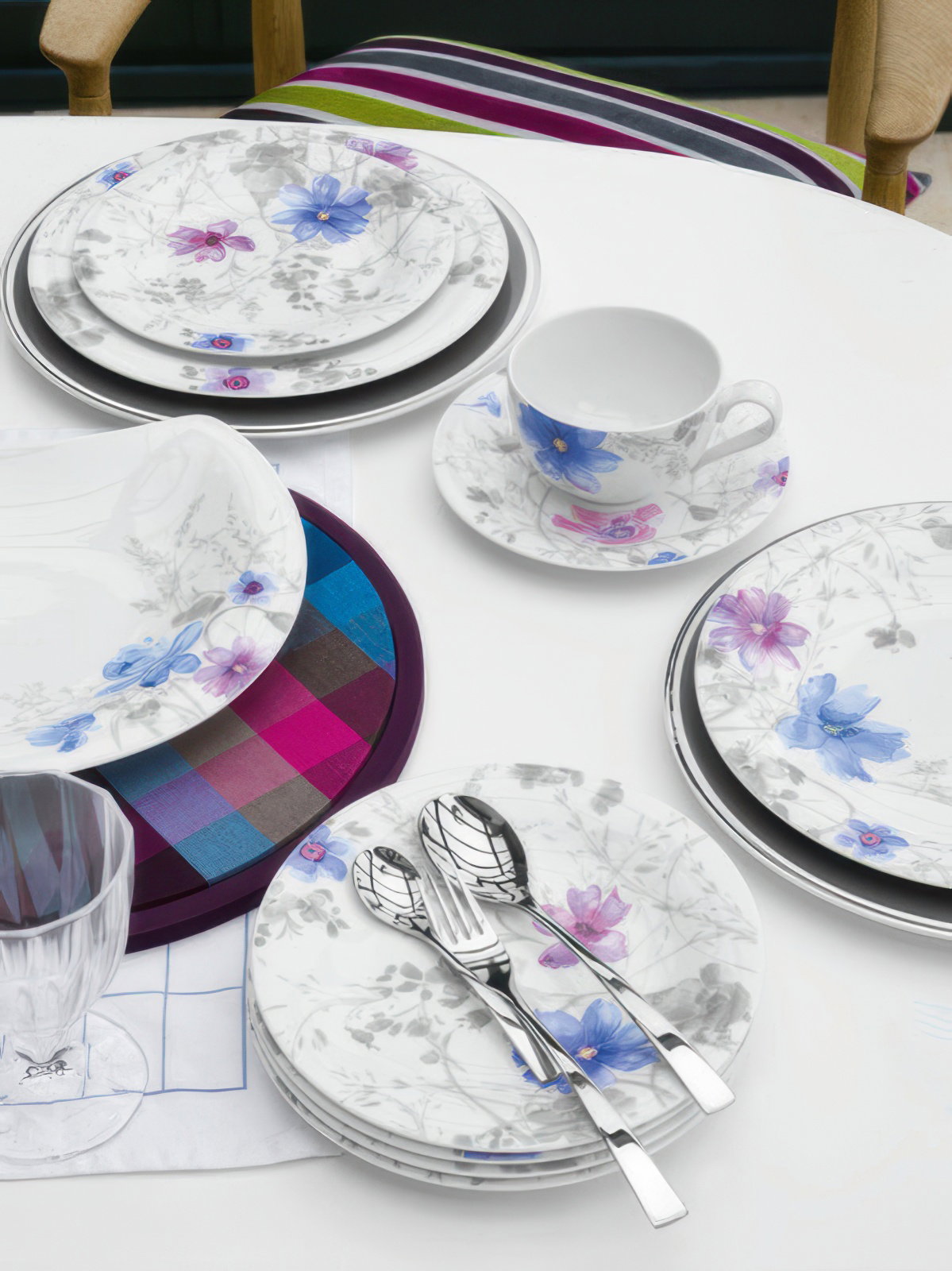 Ein Tisch mit Geschirr: Teller, Tassen und Besteck mit floralen Mustern auf einem weißen Tisch. Im Hintergrund ein kariertes Platzset.