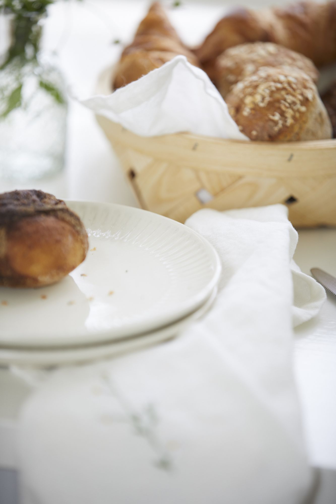 Ein Teller mit einem Brötchen steht neben einem Korb mit verschiedenen Backwaren und einer Serviette auf einem Tisch.