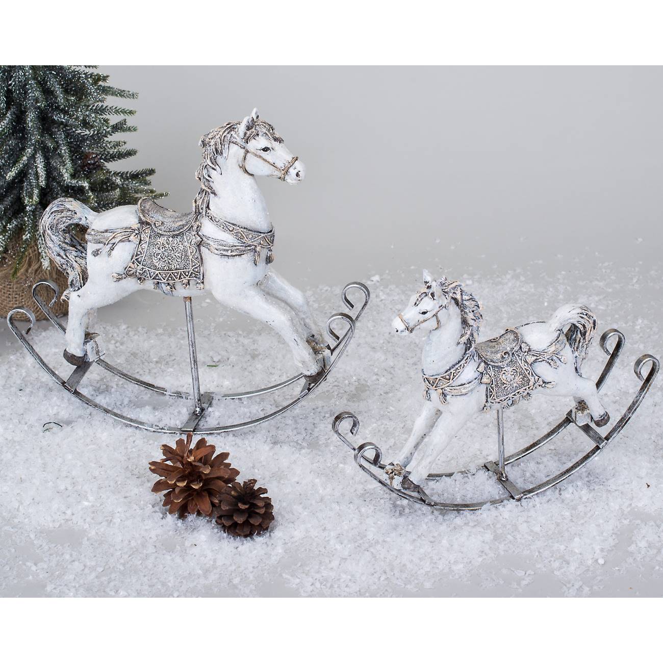 Zwei dekorative Schaukelpferde aus weißem Material mit silbernen Verzierungen, umgeben von Kunstschnee und Tannenzapfen.