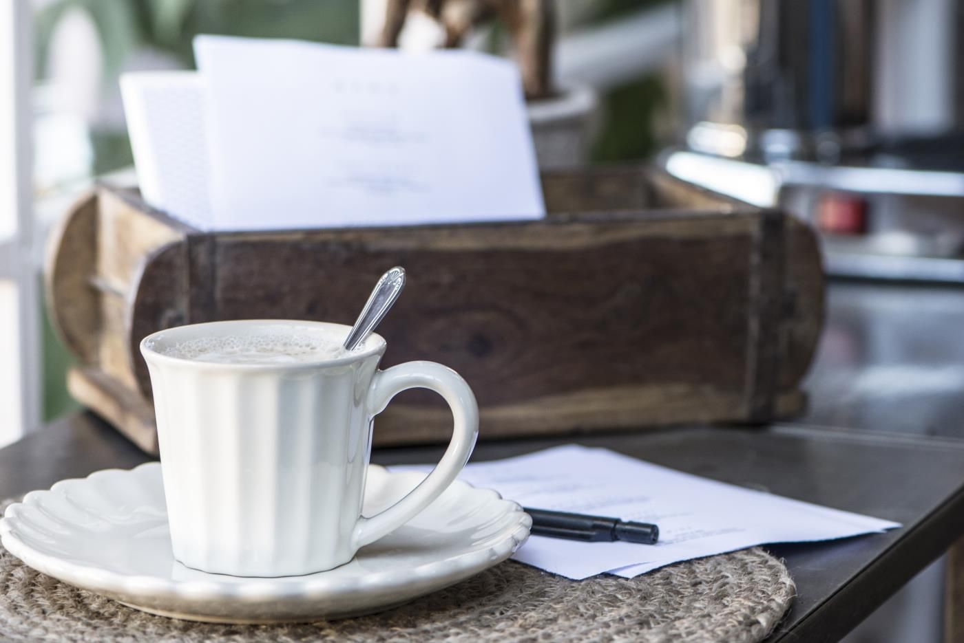 Eine Tasse Kaffee steht auf einem Unterteller. Im Hintergrund sind ein Holzbehälter und einige Blätter Papier sichtbar.