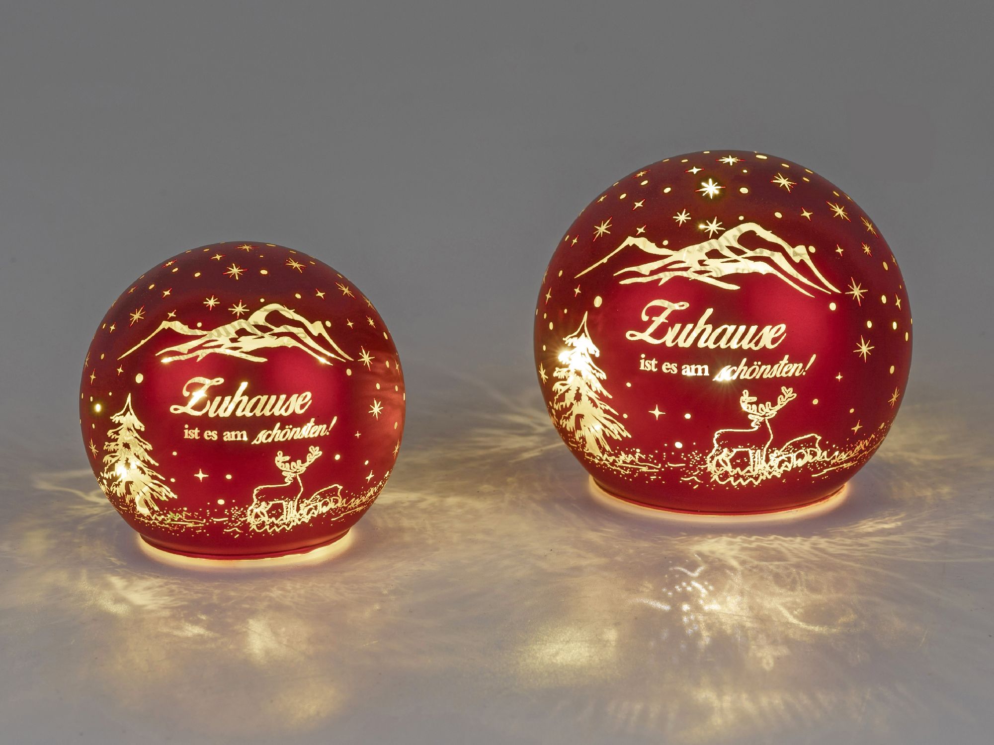 Zwei rote Glaskugeln mit Weihnachtsmotiven und dem Schriftzug „Zuhause ist es am schönsten!“ leuchte