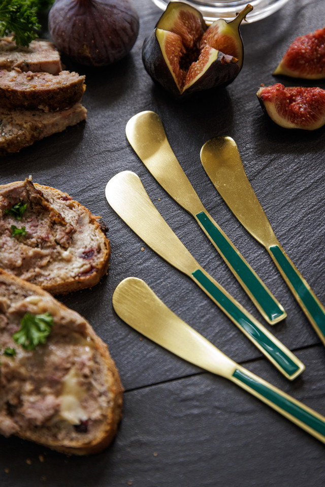 Auf einem schwarzen Untergrund liegen Scheiben von Brot mit Aufstrich, frische Feigen und goldene Messer mit grünem Griff.