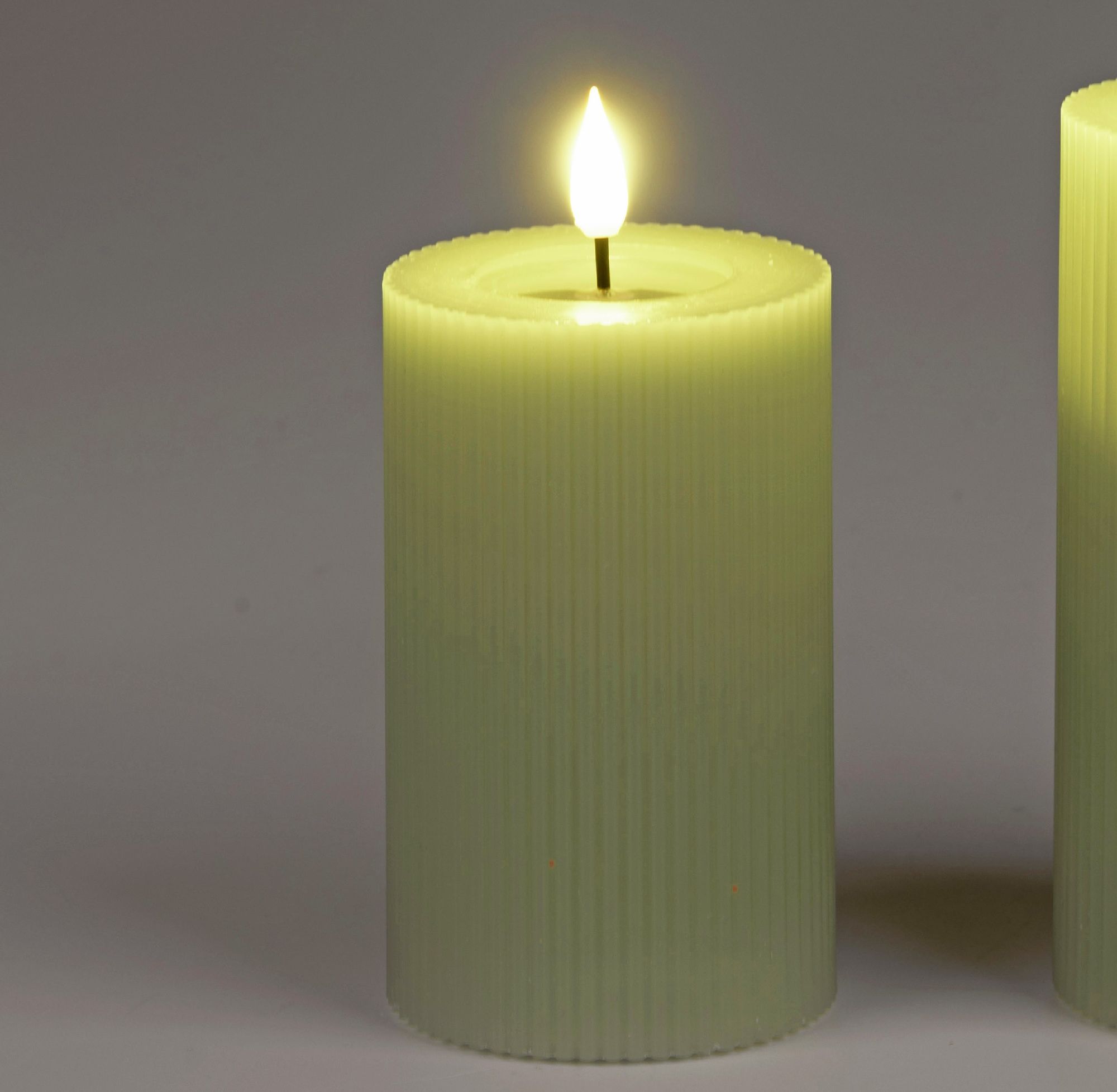 Zwei grüne, gerippte LED-Kerzen stehen nebeneinander. Eine Kerze hat eine flackernde Flamme. Hintergrund ist neutral.