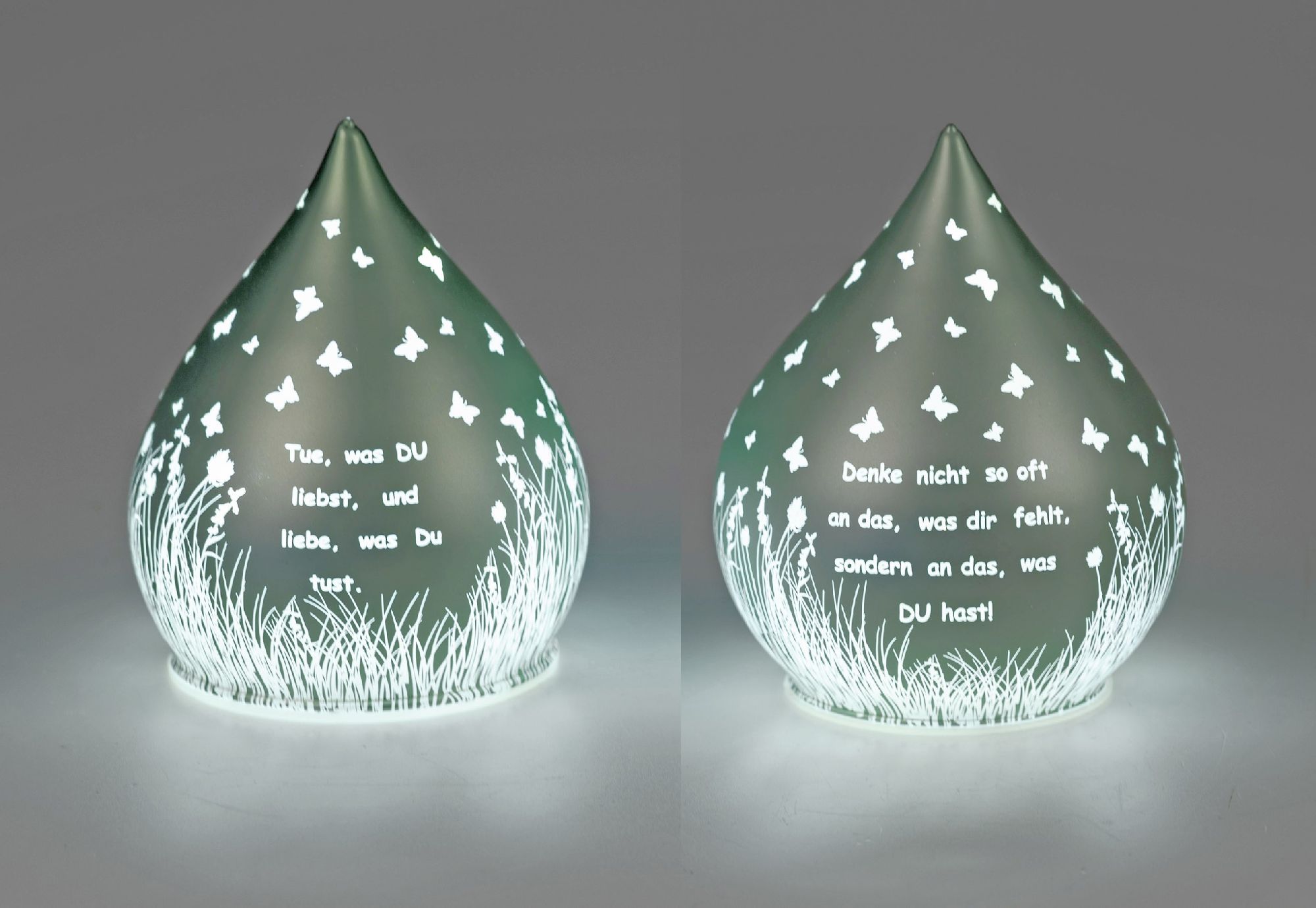 Eine gläserne, tropfenförmige Lampe mit weißen Schmetterlingen und zwei eingravierten Texten auf der Oberfläche.