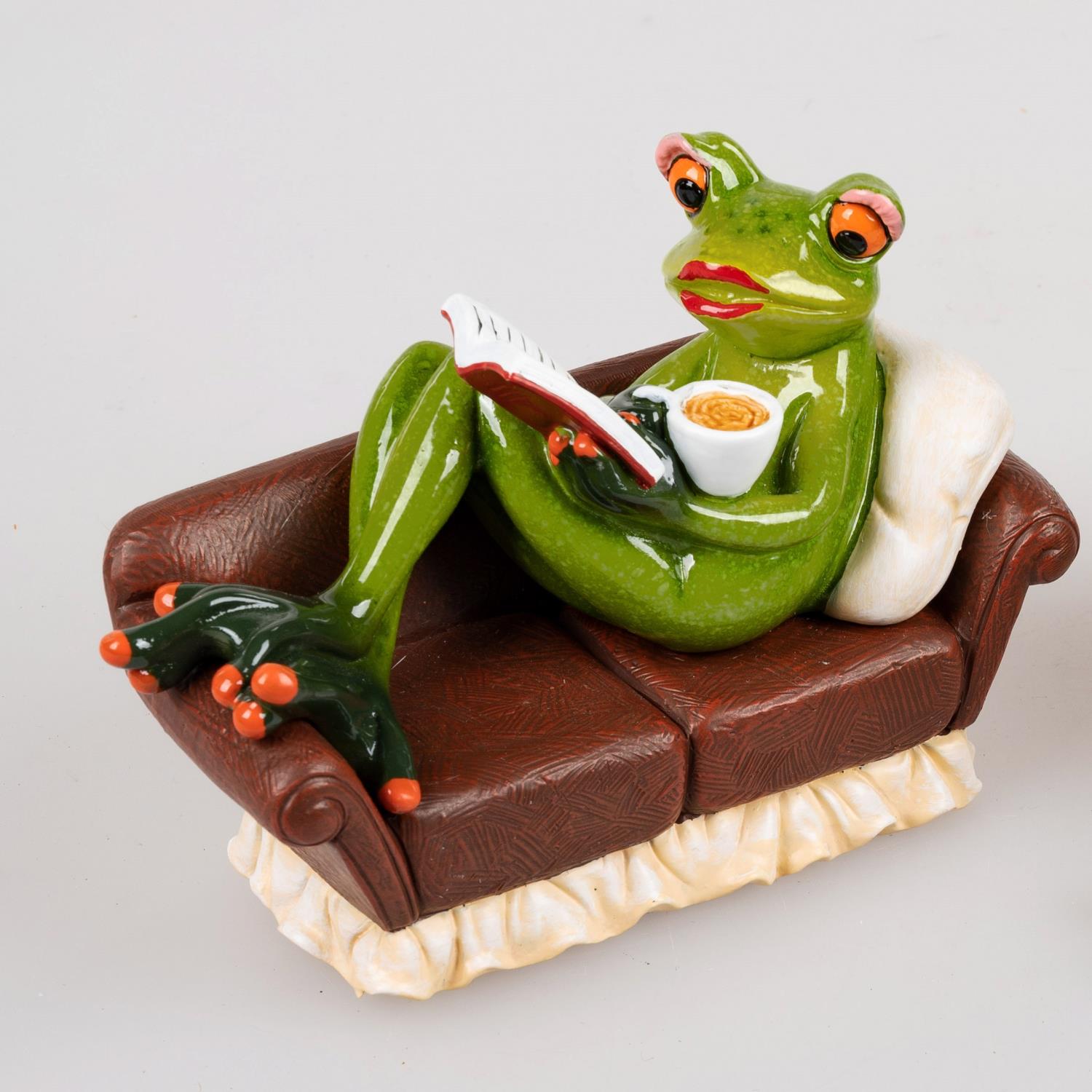 Eine grüne Froschfigur sitzt auf einem braunen Sofa, hält ein Buch in der Hand und eine Schüssel mit