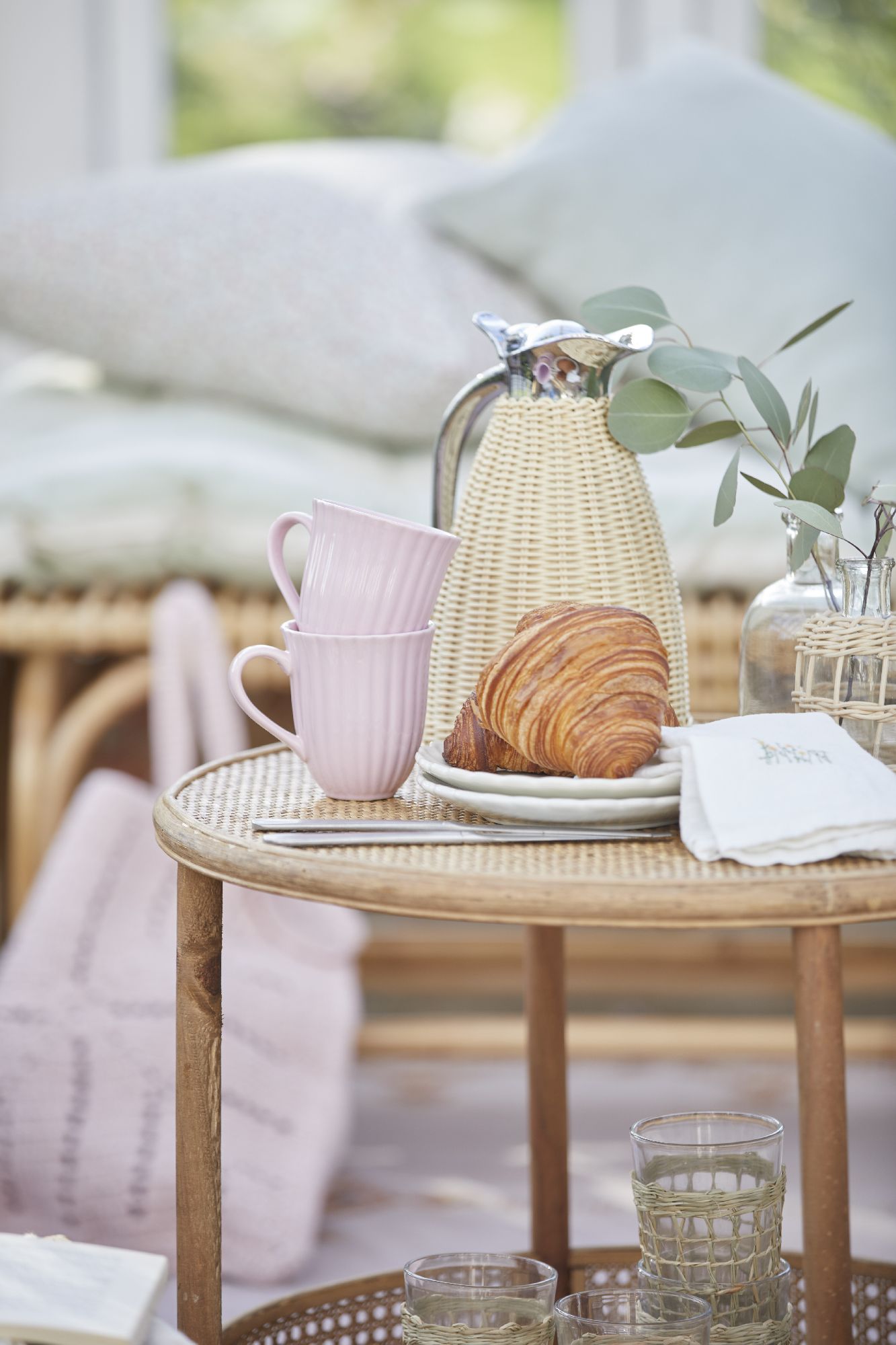 Ein Tisch mit einer Kaffeekanne, zwei rosa Tassen, einem Croissant, Gläsern und einem kleinen Zweig. Im Hintergrund Kissen.