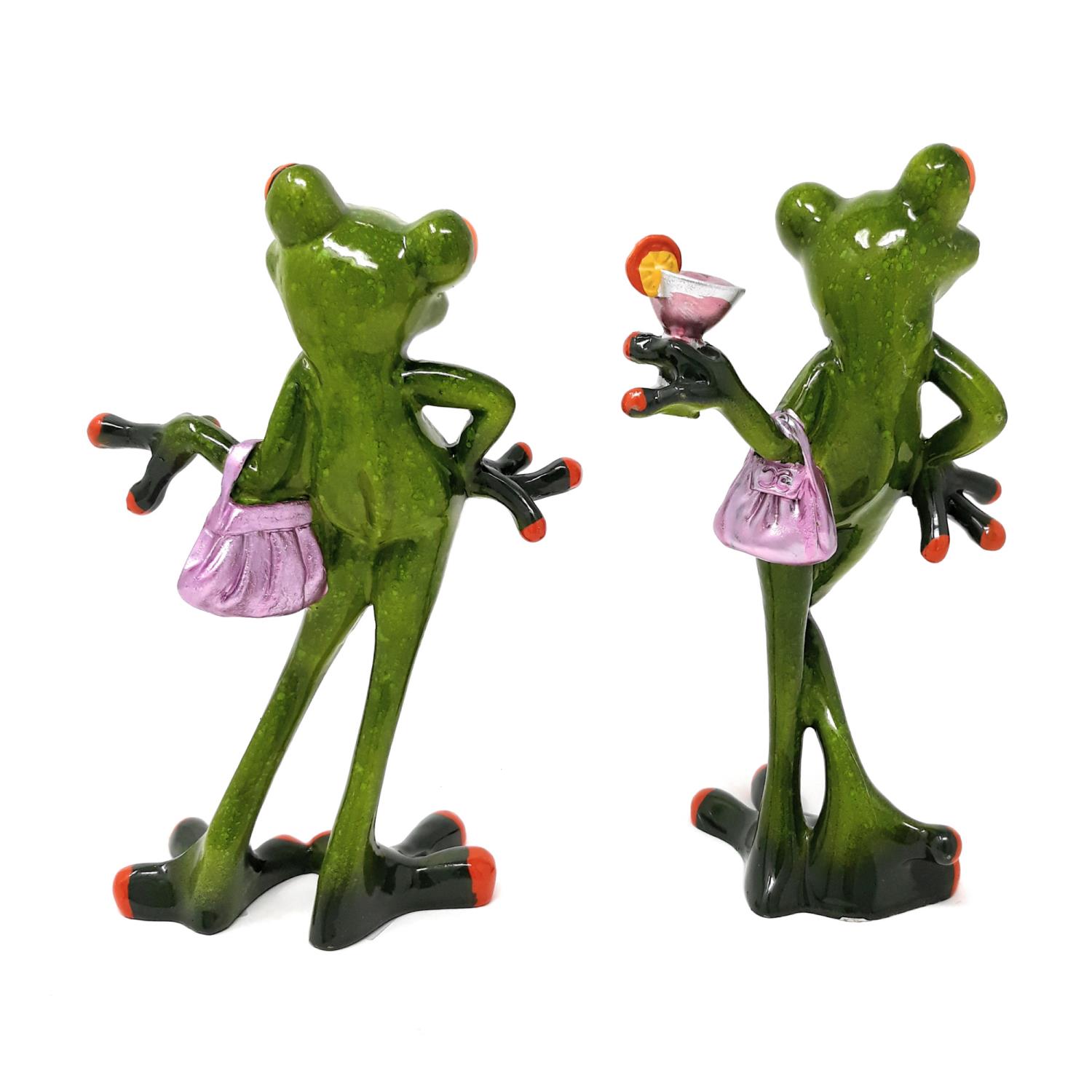 Zwei grüne Froschfiguren stehen Rücken an Rücken. Eine hält eine pinke Handtasche, die andere ein Getränk mit einer Zitrone.