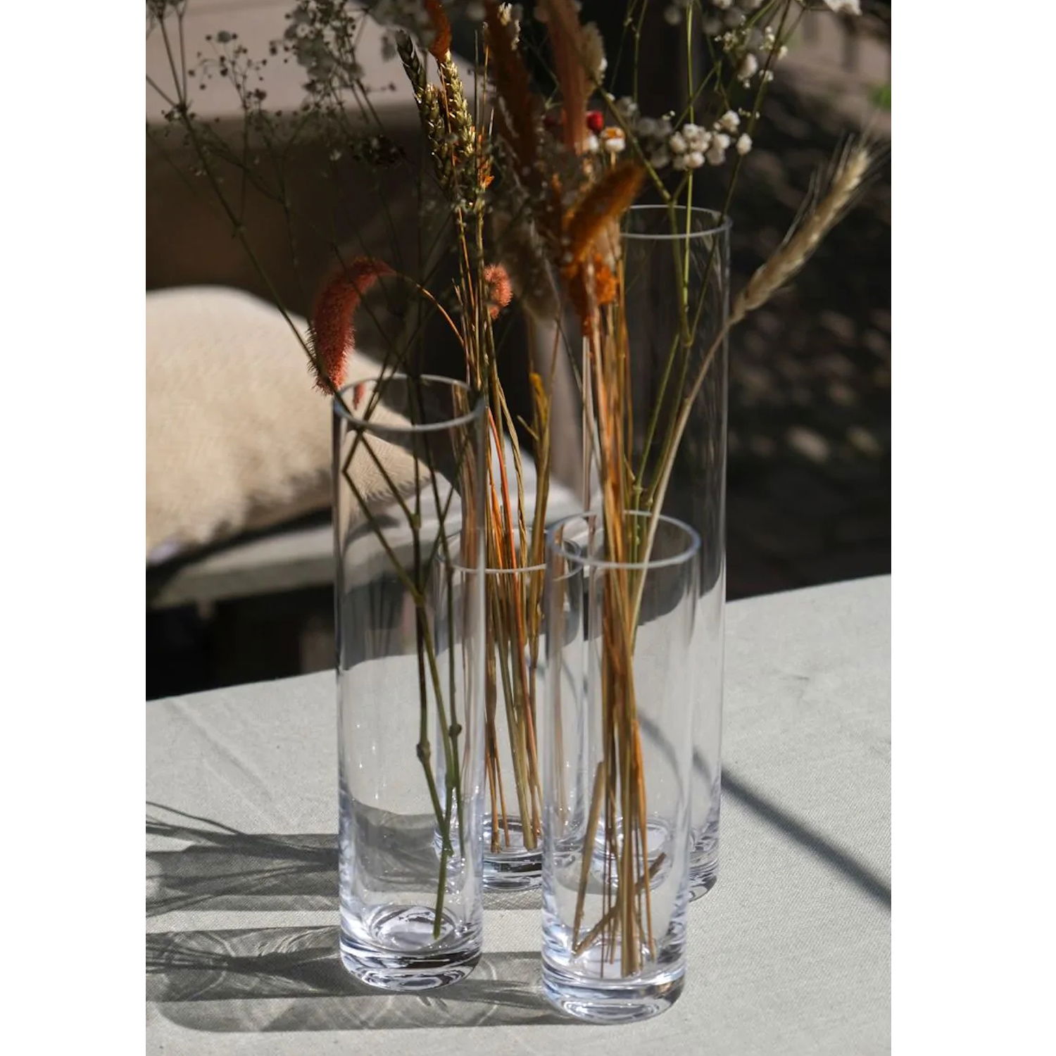 Drei hohe, klare Glasvasen mit getrockneten Blumen und Gräsern stehen auf einem Tisch. Im Hintergrund ist ein Kissen sichtbar.