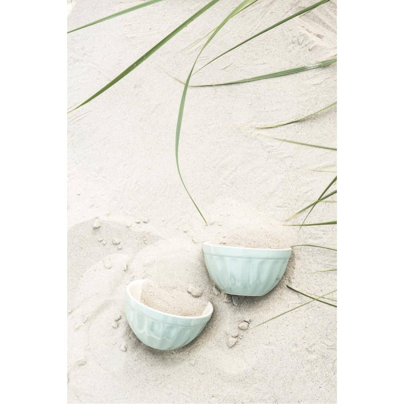 Zwei helle Schalen mit Sand stehen auf hellem Sandboden, umgeben von einigen grünen Grashalmen.