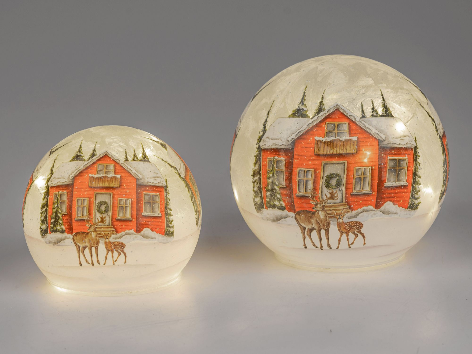 Zwei Schneekugeln mit einer roten Hütte im Schnee, umgeben von Tannen und zwei Rehen. Die Kugeln sin