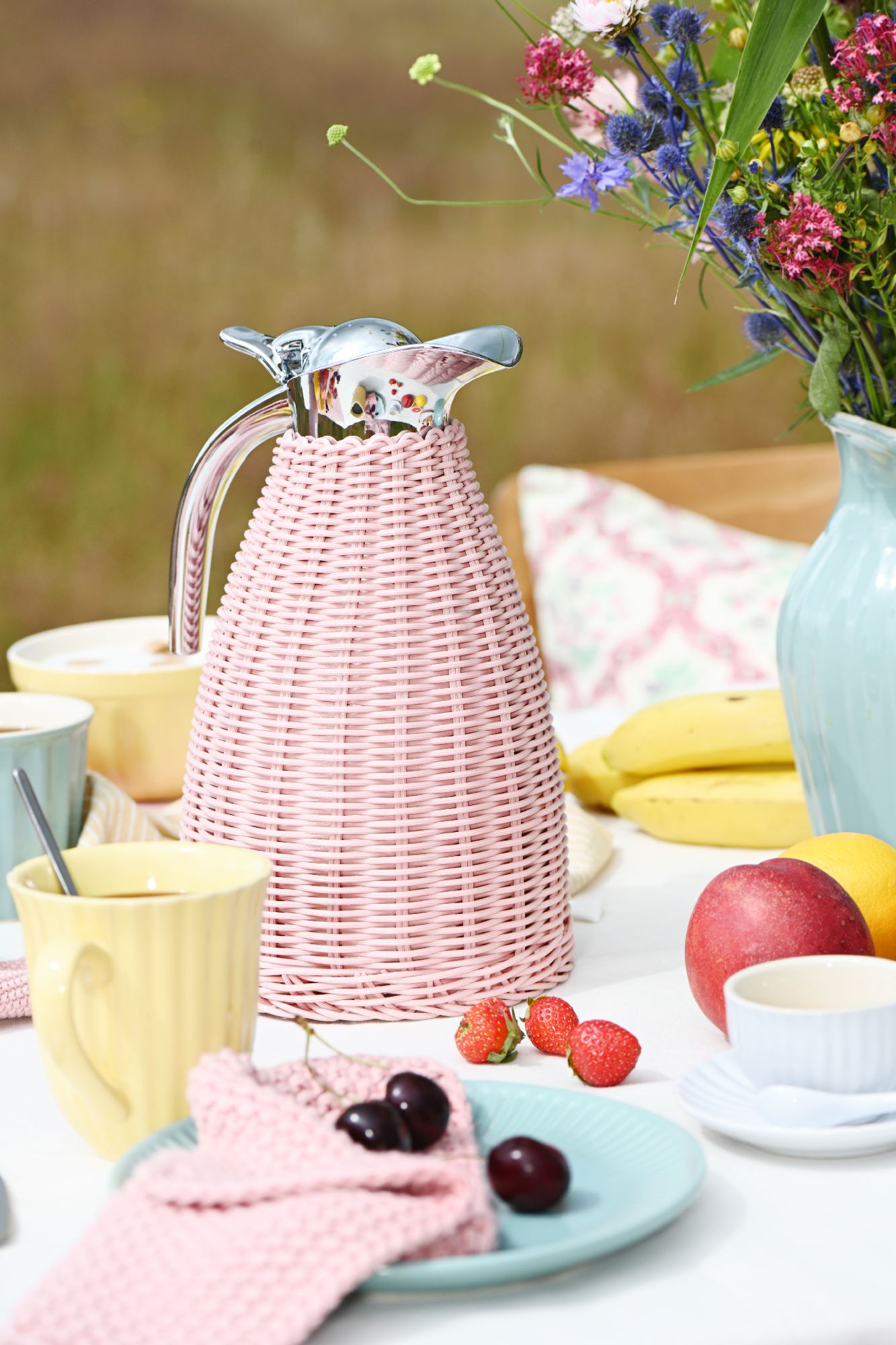 Ein Tischgedeck mit einer pinken Kanne, Tassen, Obst und einer Blumenvase in einer natürlichen Umgebung.