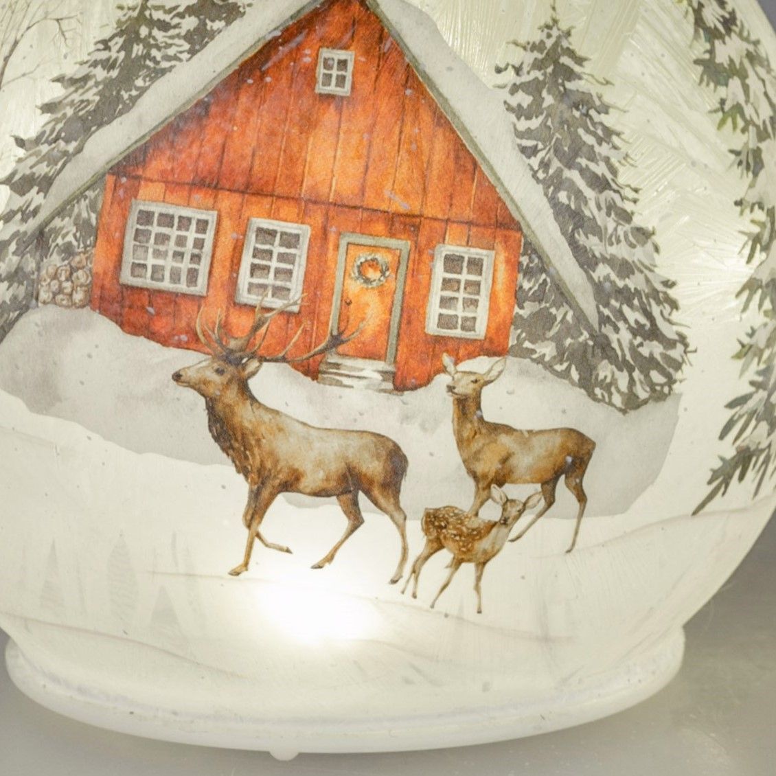 Eine rot gestrichene Hütte im Schnee, umgeben von Tannen, mit zwei Rehen und einem Rehkitz im Vorder