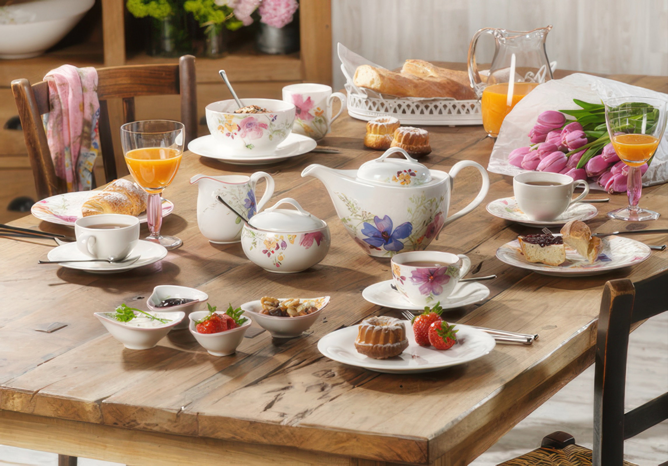 Ein gedeckter Tisch mit Teekanne, Tassen, Gebäck, Obst und Blumenarrangement. Getränke in Gläsern sind ebenfalls sichtbar.