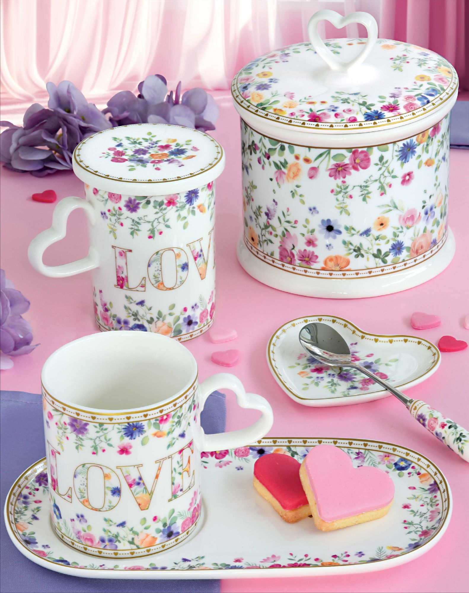 Tasse und Dose mit Blumenmuster, Herzgriff und dem Wort "LOVE". Dazu Herzplätzchen auf einem Teller. Rosa und lila Hintergrund.