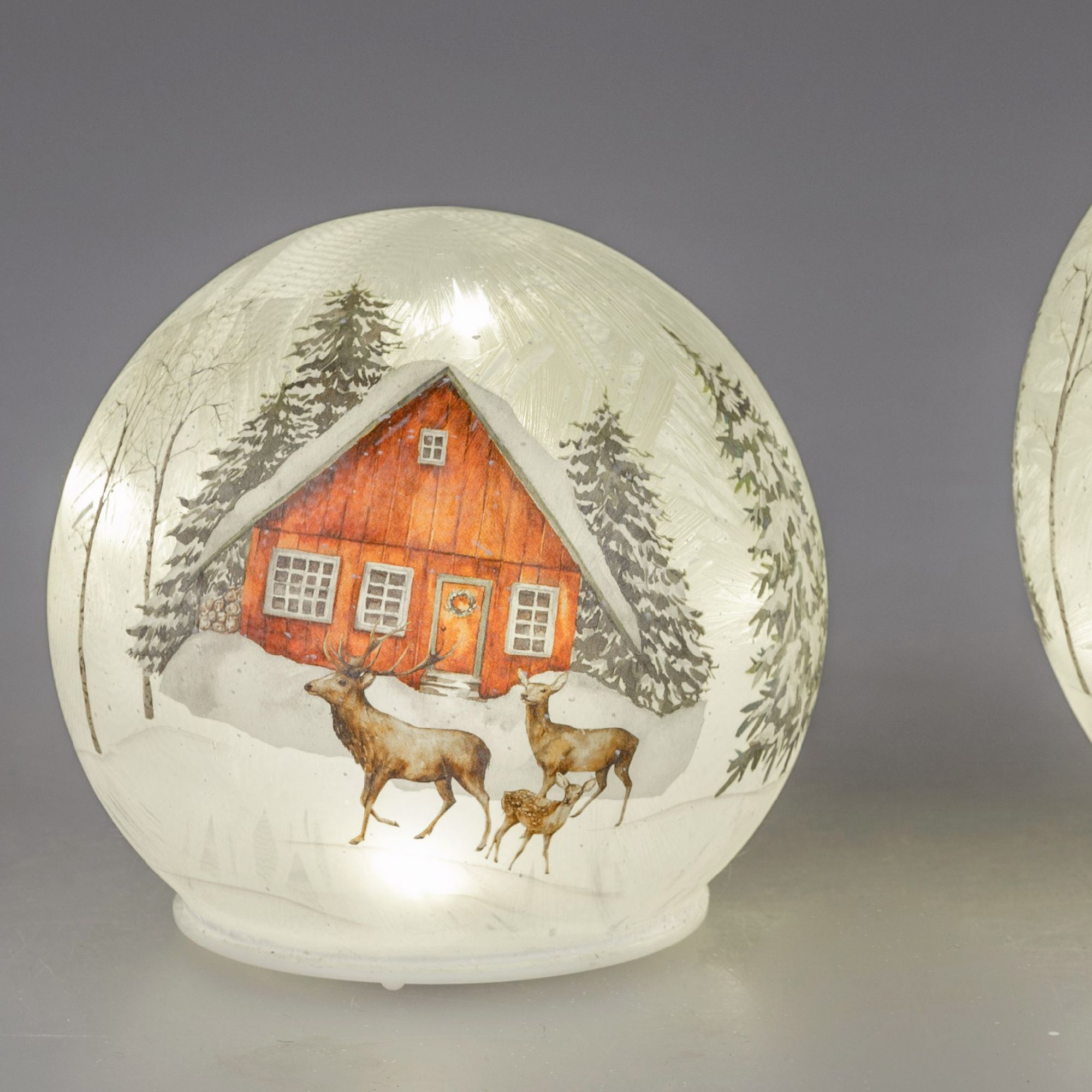 Eine Schneekugel zeigt ein rotes Holzhaus im Schnee, umgeben von Tannenbäumen und drei Rehen.