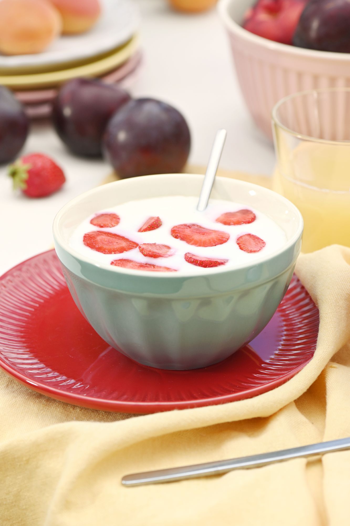Eine Schüssel mit Joghurt und Erdbeerscheiben steht auf einem roten Teller. Im Hintergrund sind Obst und ein Glas Saft sichtbar.