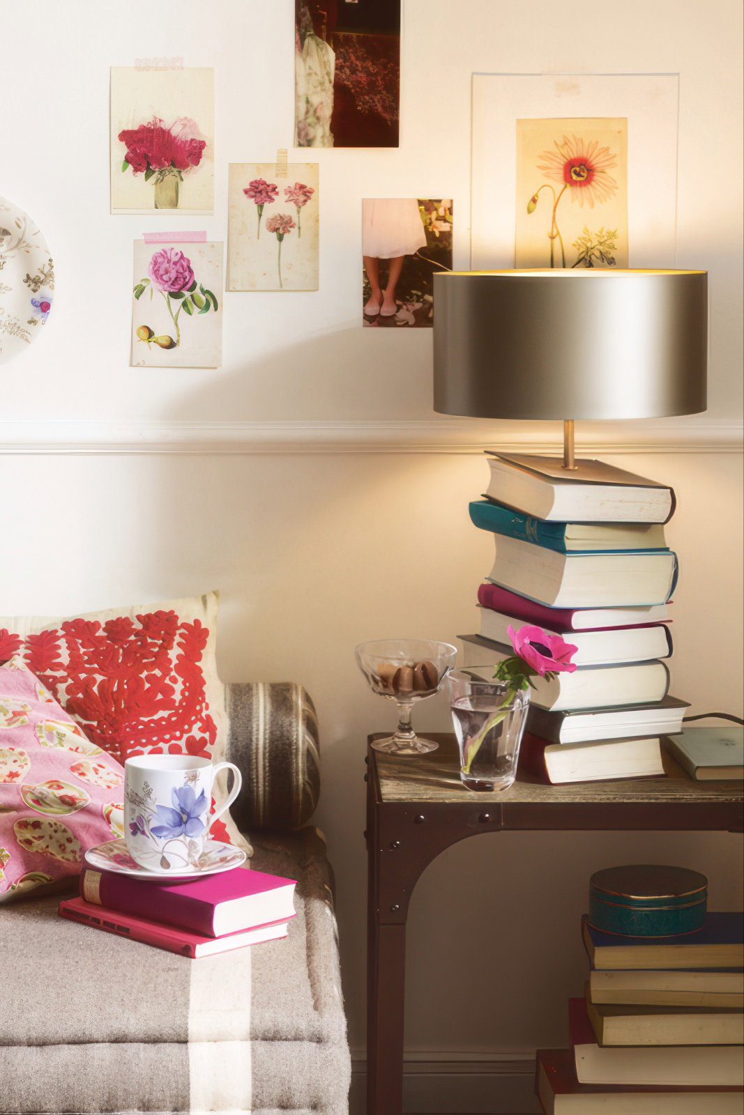 Ein gemütlicher Raum mit einem Sofa, einem Tisch mit Büchern, einer Lampe und Dekoration an der Wand. Eine Tasse und eine Blume sind sichtbar.