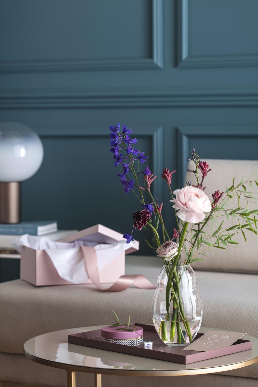 Vase mit bunten Blumen auf einem Tisch, daneben ein Geschenk in einer offenen Schachtel und ein kleiner runder Behälter.