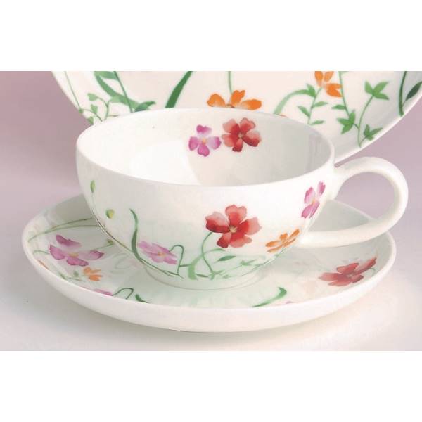 Eine weiße Tasse mit bunten Blumenmustern steht auf einer Untertasse. Im Hintergrund ist ein flacher Teller mit ähnlichem Design sichtbar.