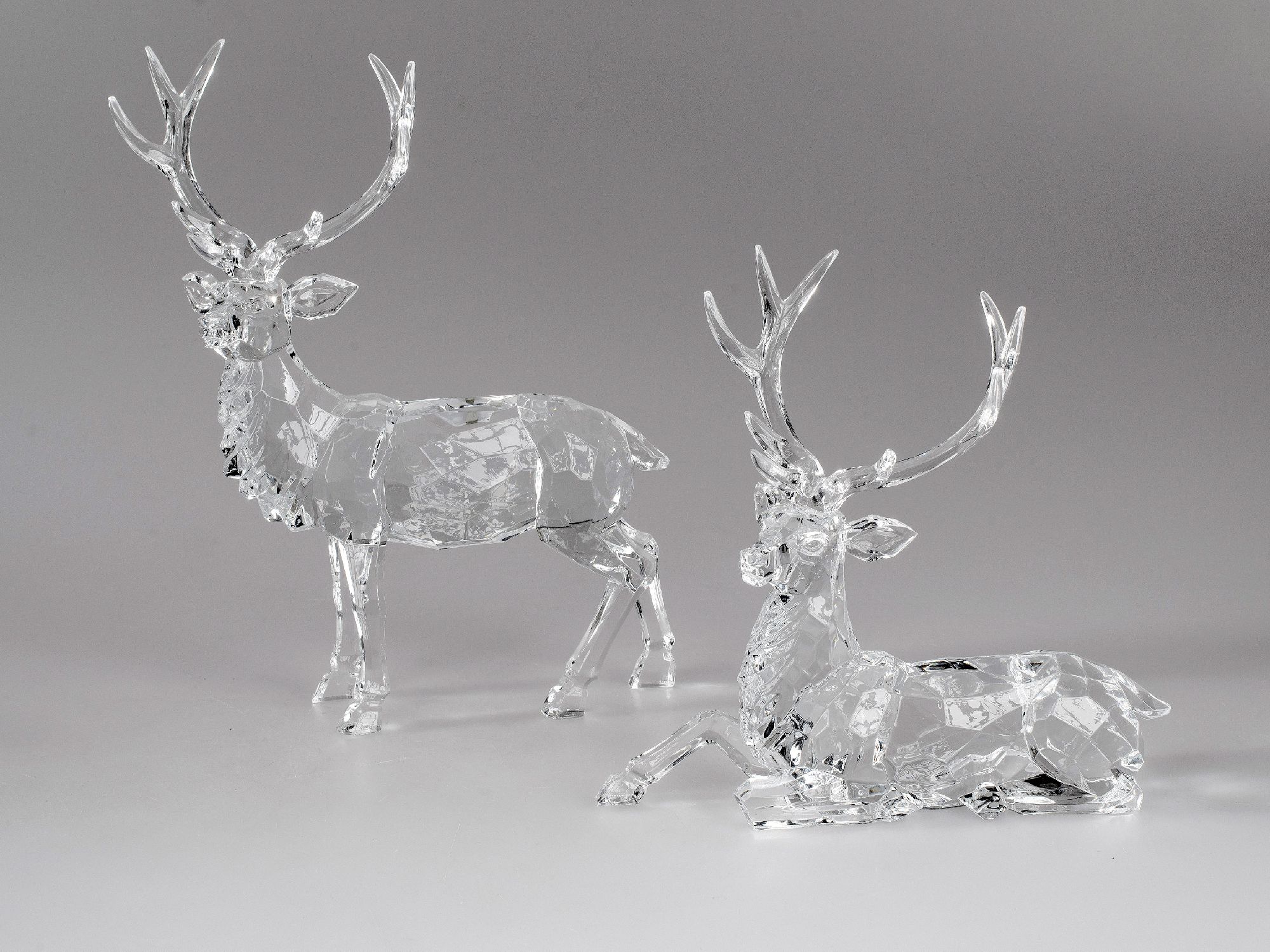 Zwei durchsichtige, gläserne Rehe stehen und liegen auf einem grauen Hintergrund. Die Figuren sind d