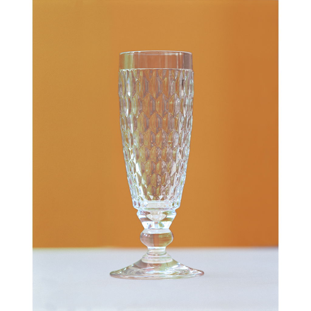 Ein hohes, facettiertes Glas mit einem stabilen Fuß steht auf einem Tisch vor einem orangefarbenen Hintergrund.