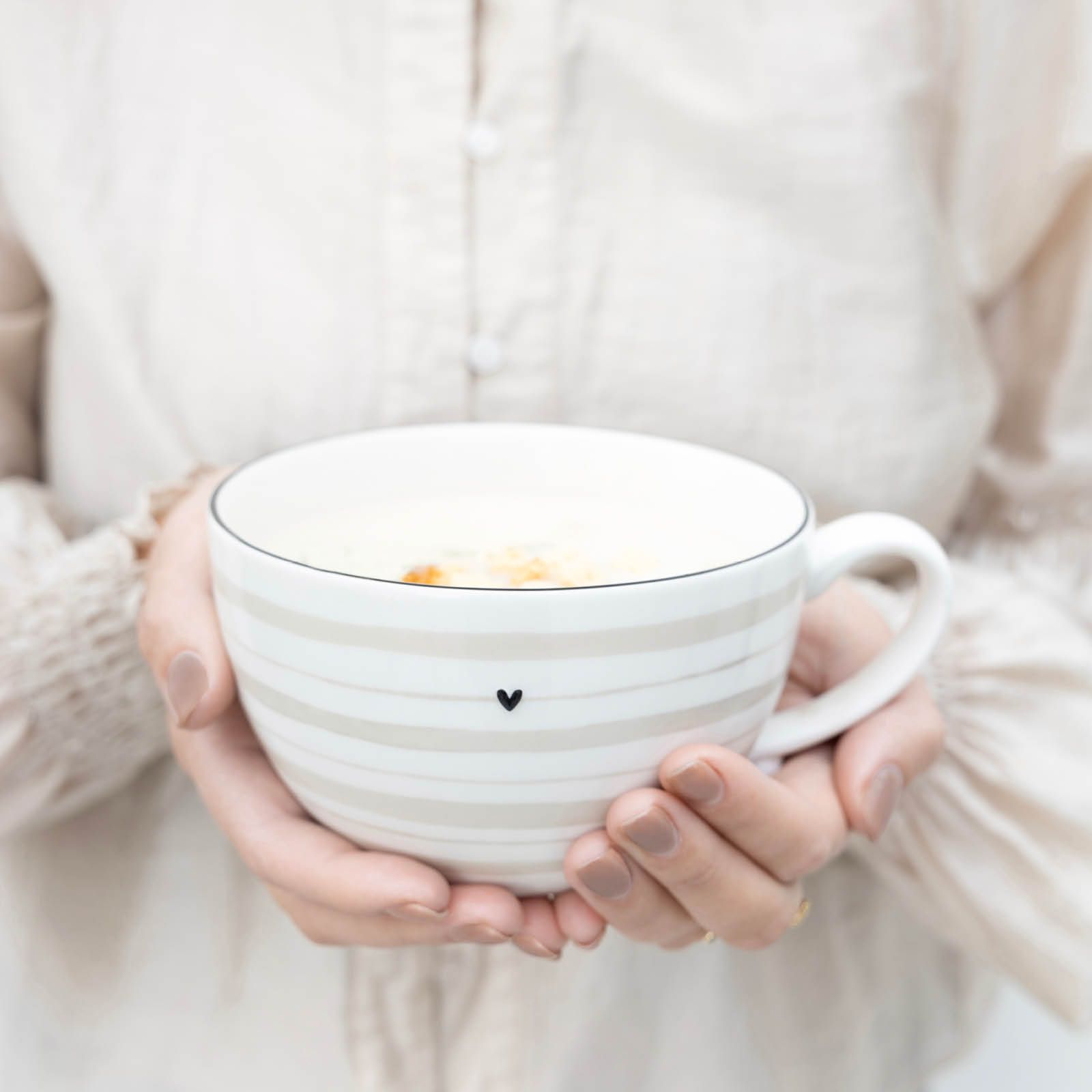 Eine Person hält eine große, gestreifte Tasse mit einem kleinen Herzsymbol. Die Tasse ist gefüllt und hat einen Henkel.
