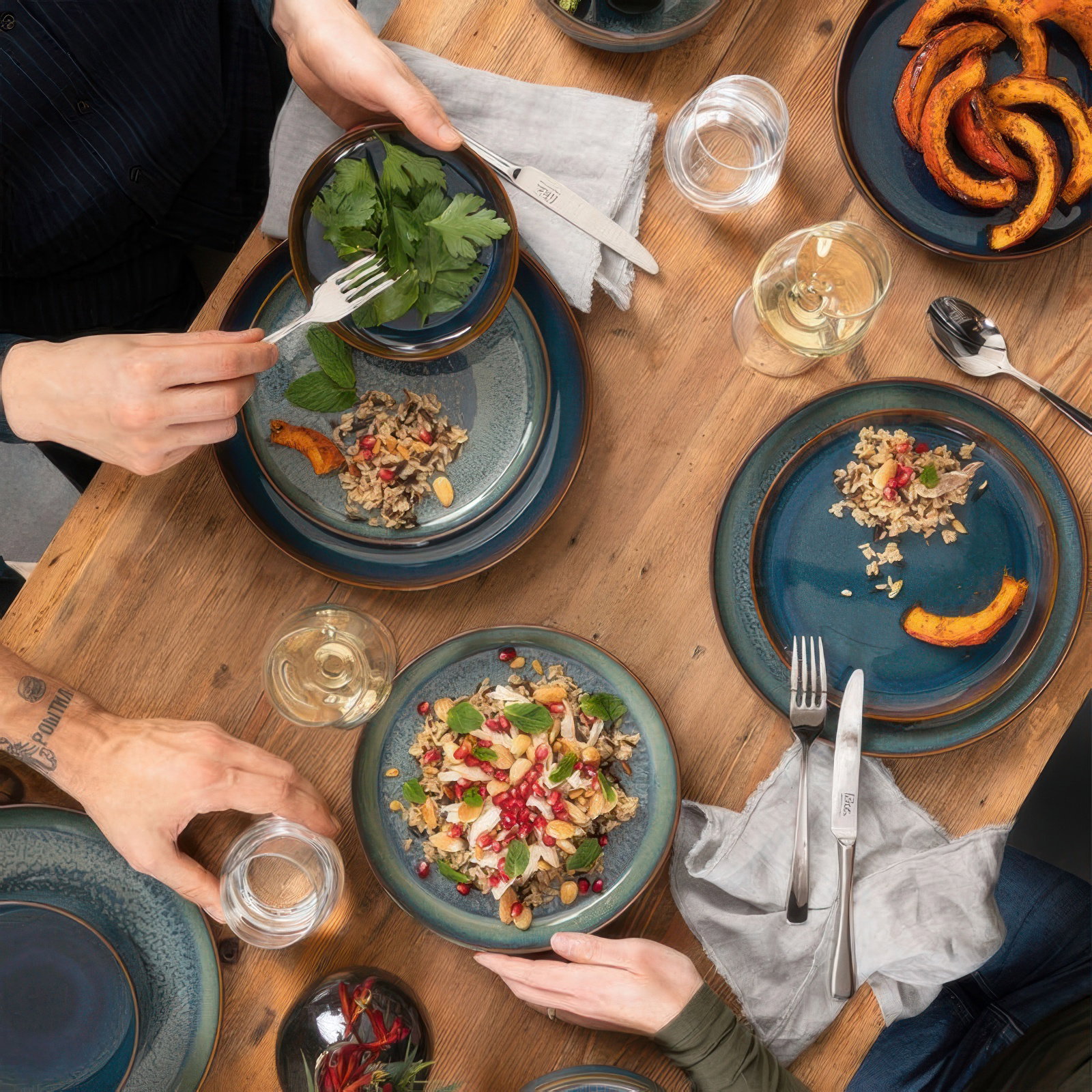 Tisch mit mehreren Tellern, Salat, Kräutern, Kürbisstücken und Gläsern. Hände greifen nach Essen und Getränken. Holzoberfläche.