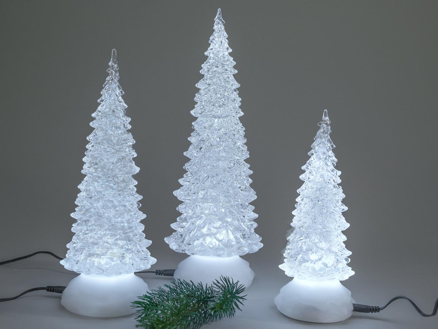 Drei leuchtende, gläserne Weihnachtsbäume in verschiedenen Größen stehen auf einer weißen Basis, umgeben von einem kleinen grünen Zweig.