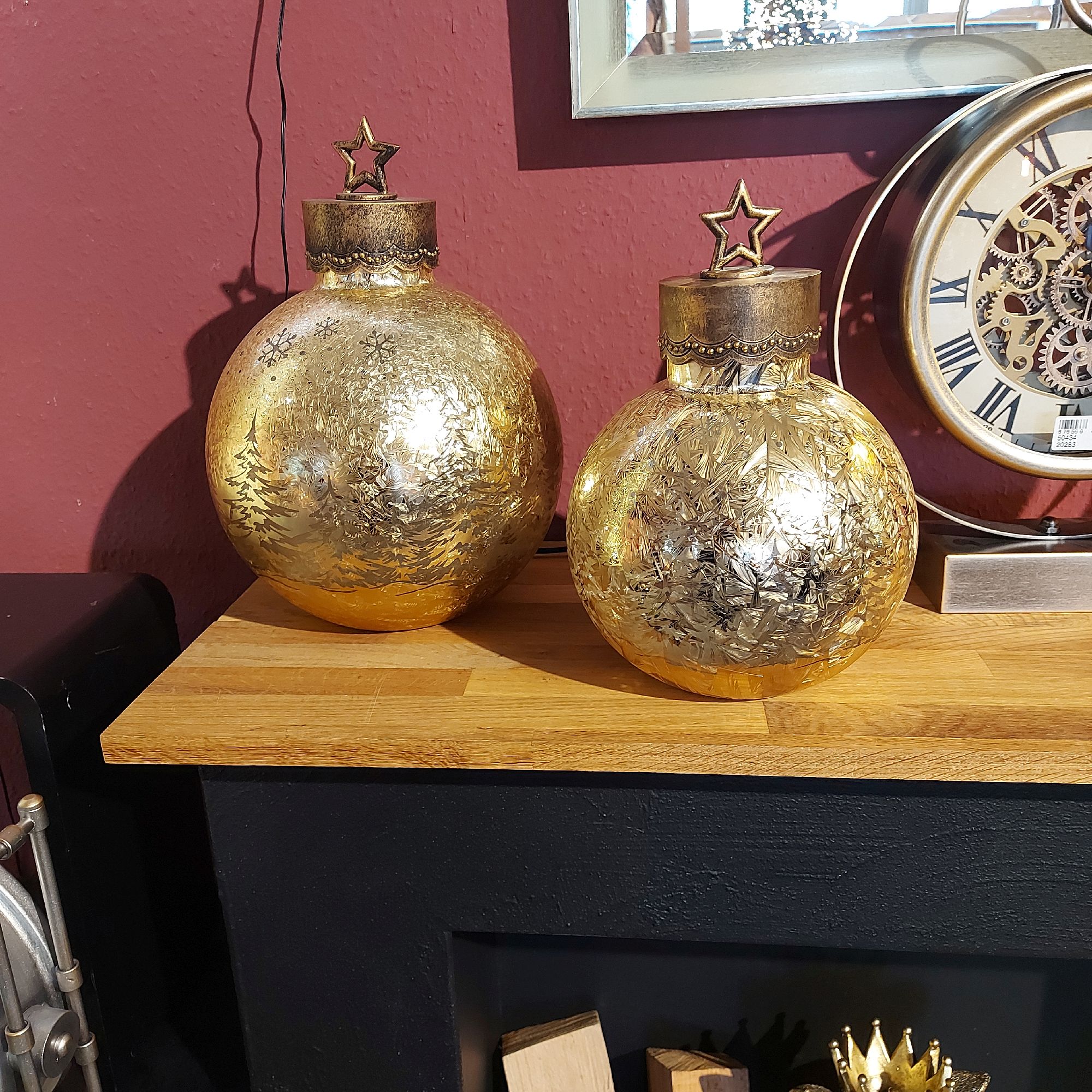 Zwei goldene Dekokugeln mit Sternenaufsatz stehen auf einem Holzregal vor einer roten Wand. Im Hinte