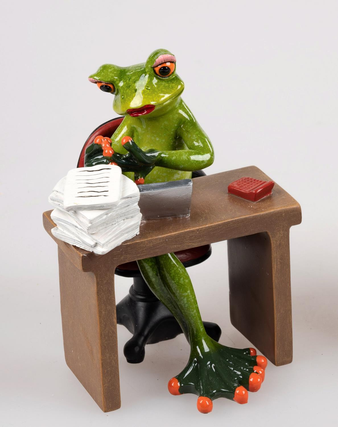 Eine grüne Froschfigur sitzt an einem Schreibtisch, hält einen Stift und schaut auf einen Stapel Pap