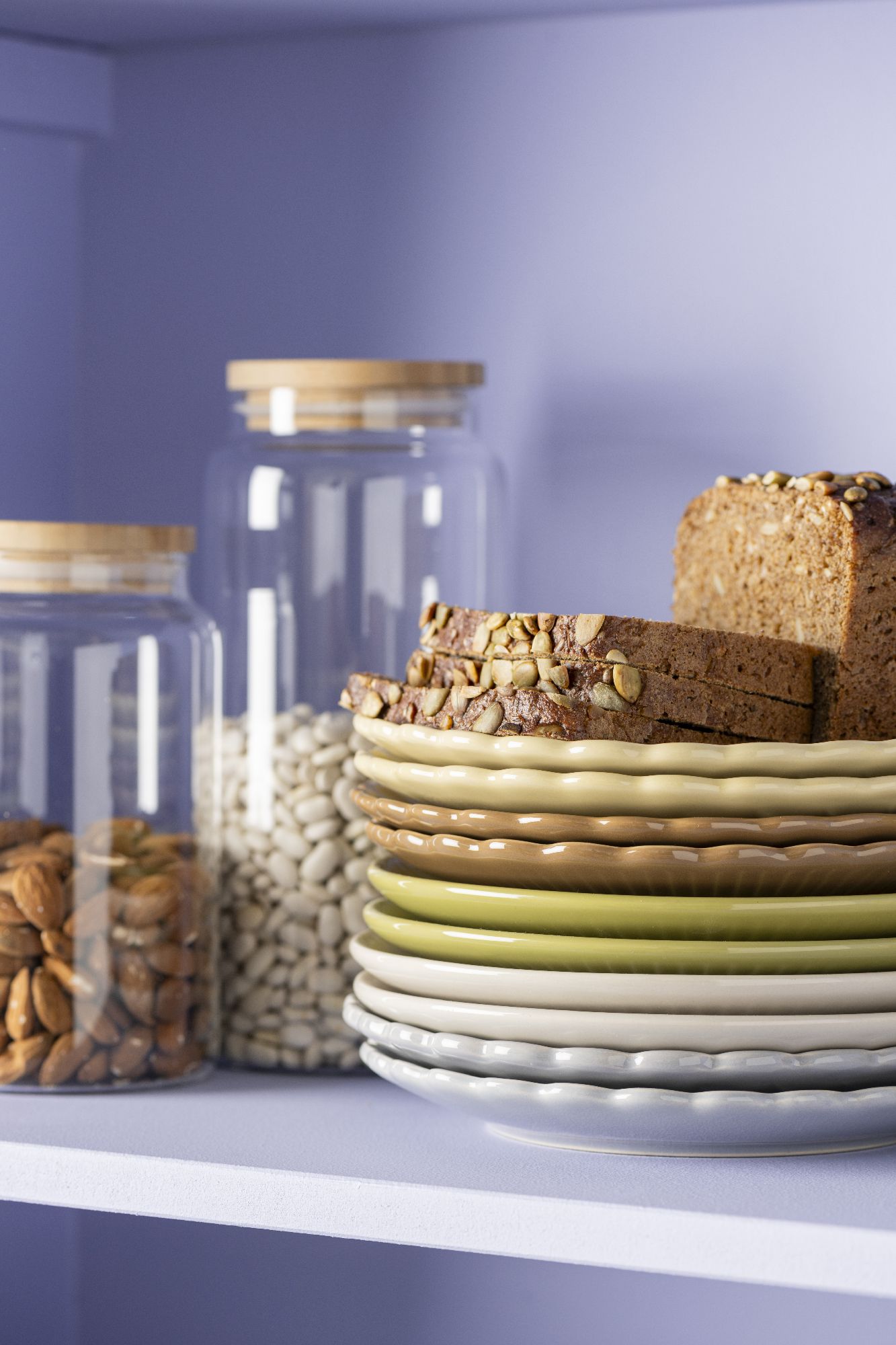 Gestapelte Teller in verschiedenen Farben, daneben Gläser mit Nüssen und Bohnen sowie ein Stück Brot. Hintergrund in lila.