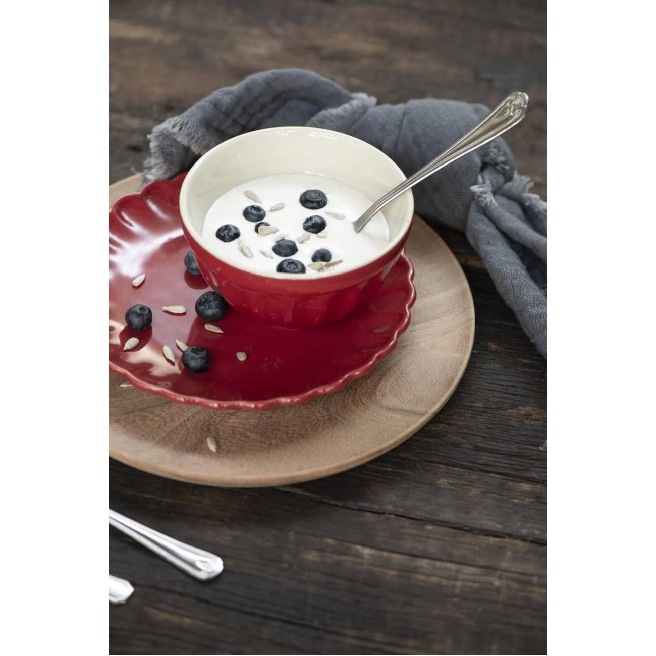 Eine rote Schüssel mit Joghurt, garniert mit Heidelbeeren und Sonnenblumenkernen, steht auf einem Holzbrett. Ein Löffel liegt daneben.