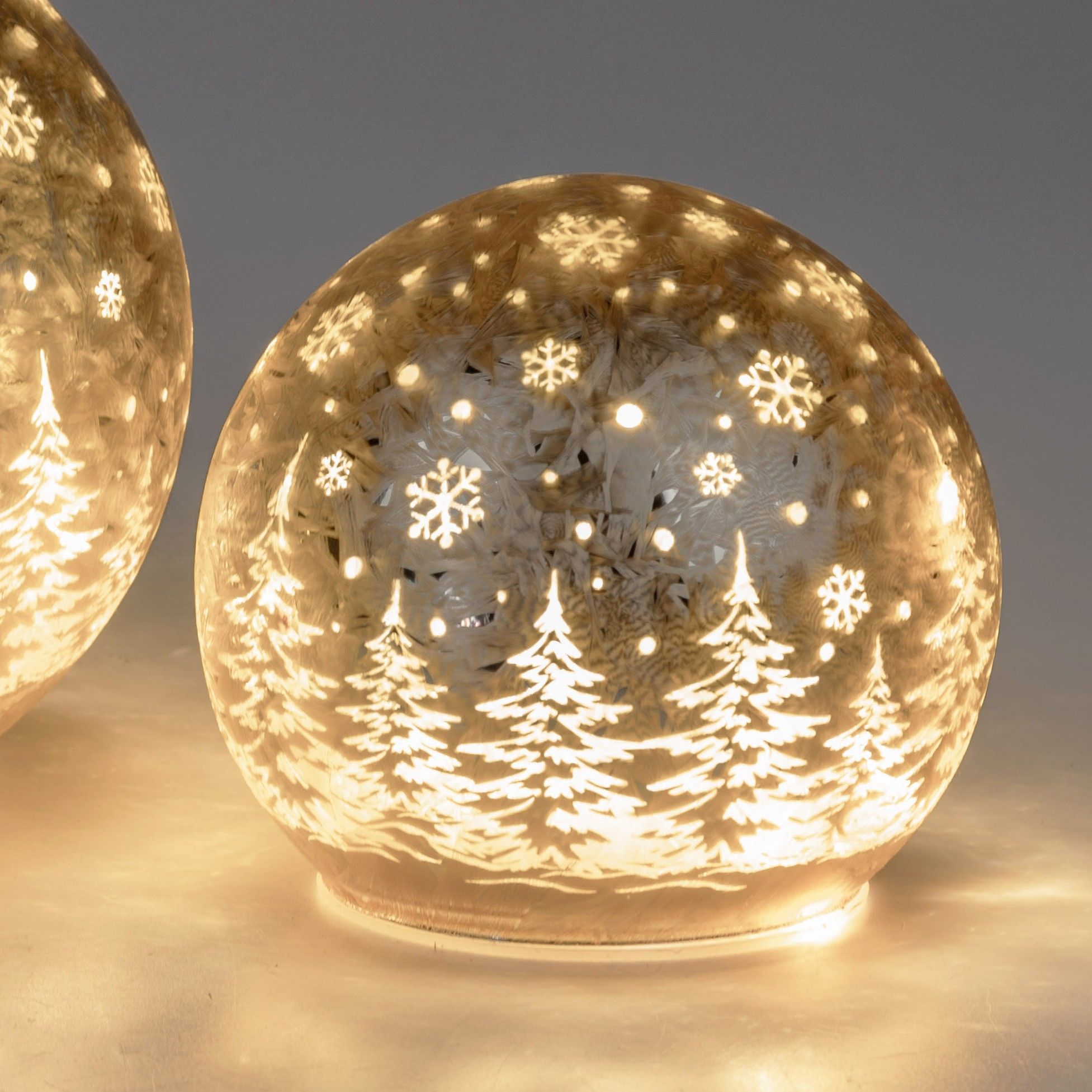 Zwei gläserne Kugeln mit beleuchteten, winterlichen Motiven von Tannenbäumen und Schneeflocken auf h