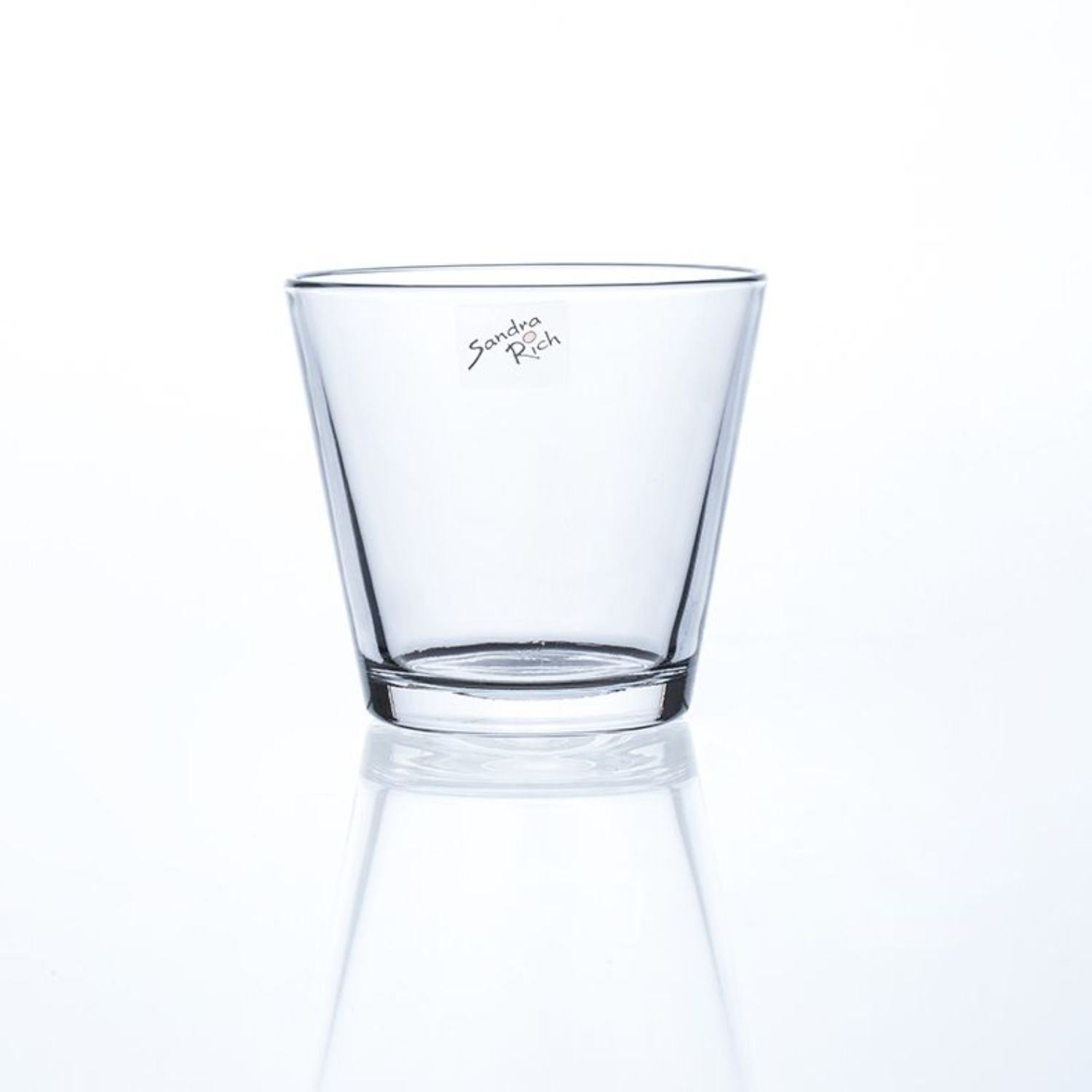 Ein transparenter, konischer Glasbecher steht auf einer reflektierenden Oberfläche. Auf dem Glas ist eine Unterschrift zu sehen.