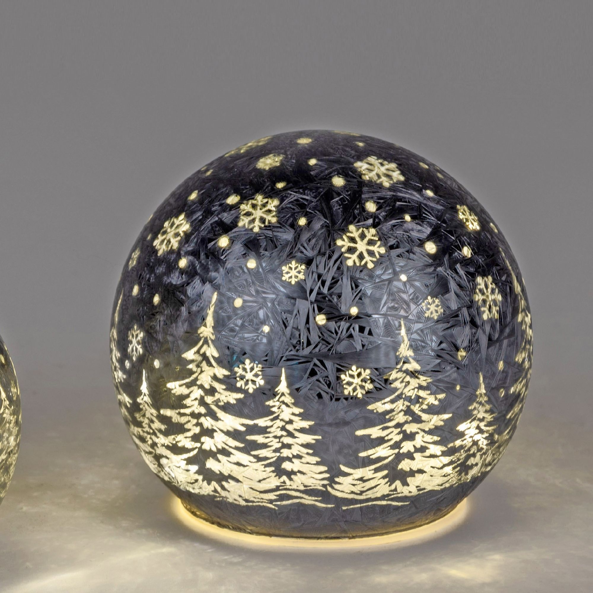 Runde, schwarze Weihnachtskugel mit weißen Schneeflocken und Tannenbäumen, die sanft beleuchtet ist.
