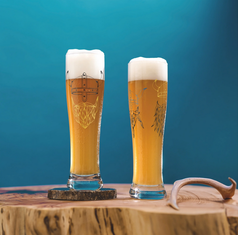 Zwei Gläser mit hellem Bier und Schaumkrone stehen auf einem Holzbrett. Im Hintergrund ist eine blaue Wand zu sehen.