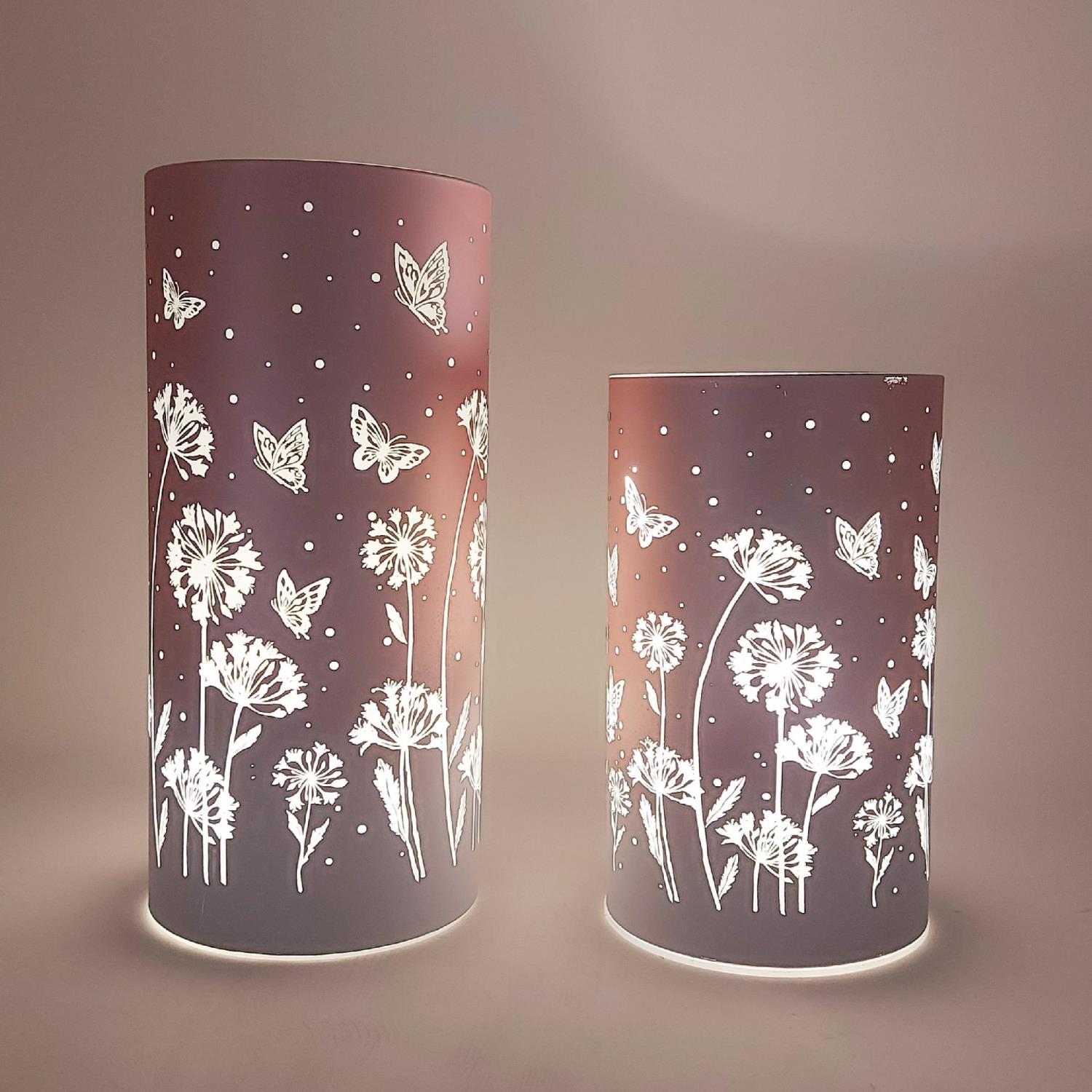 Zwei zylindrische Lampen mit floralem Muster und Schmetterlingen, beleuchtet und auf einem einfarbigen Hintergrund platziert.