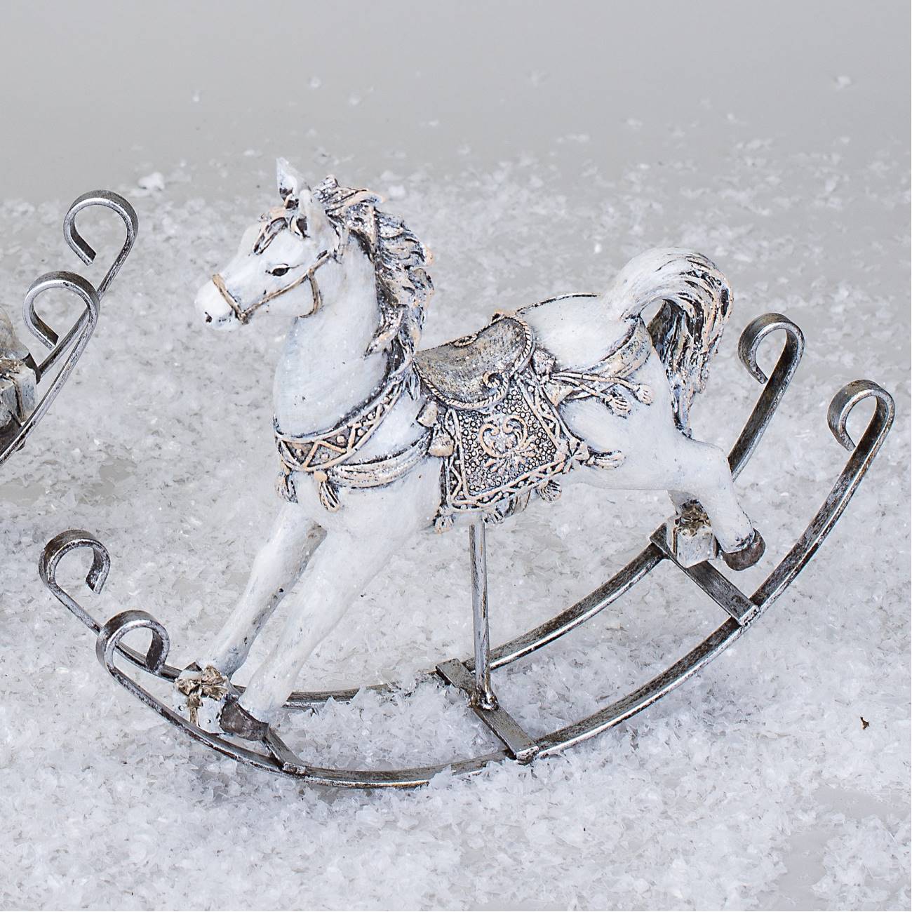 Ein weißes Schaukelpferd aus Metall mit detaillierter Verzierung steht auf einer schneebedeckten Fläche.