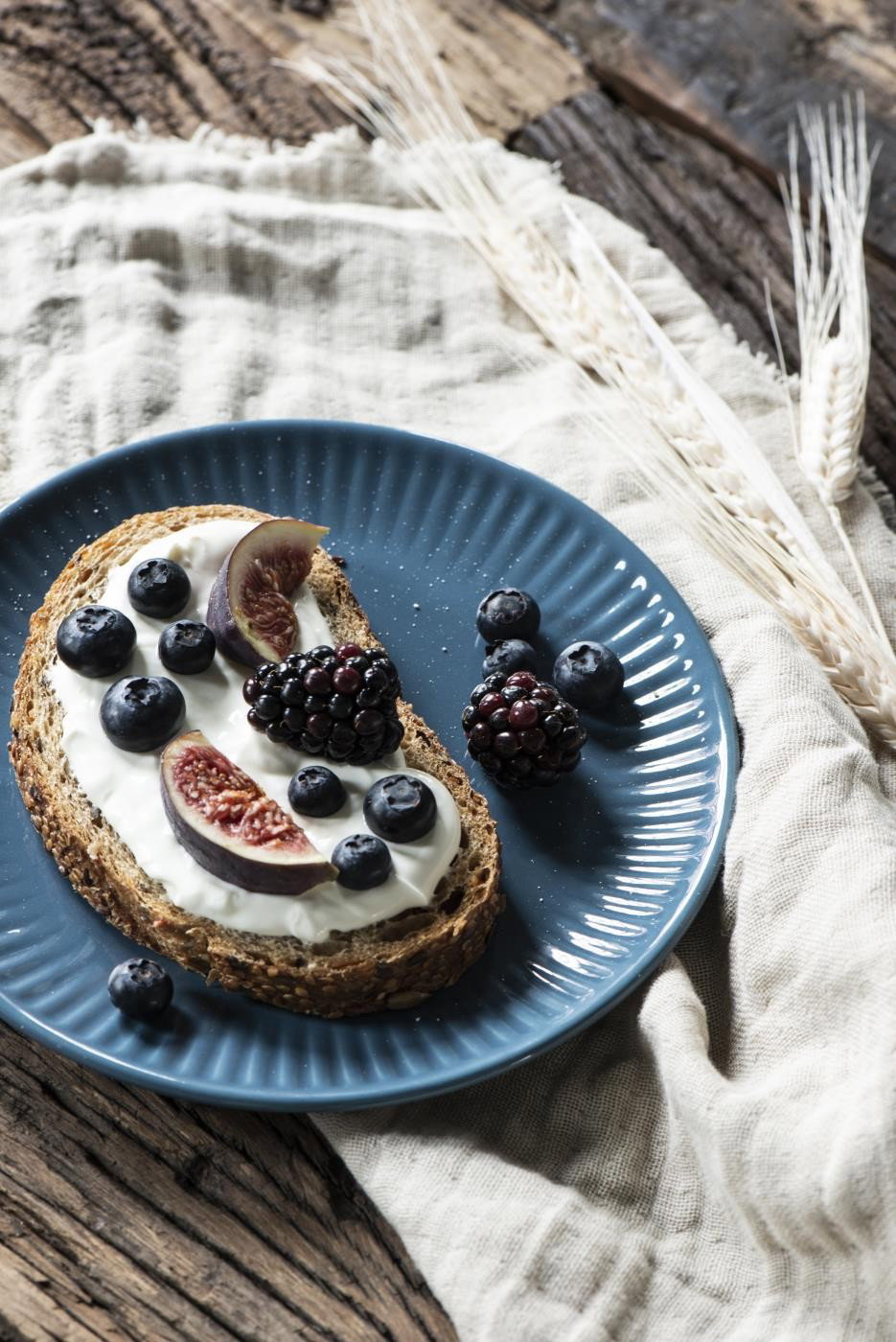 Ein Stück Vollkornbrot mit Joghurt, Feigen und verschiedenen Beeren auf einem blauen Teller, umgeben von einem hellen Tuch.