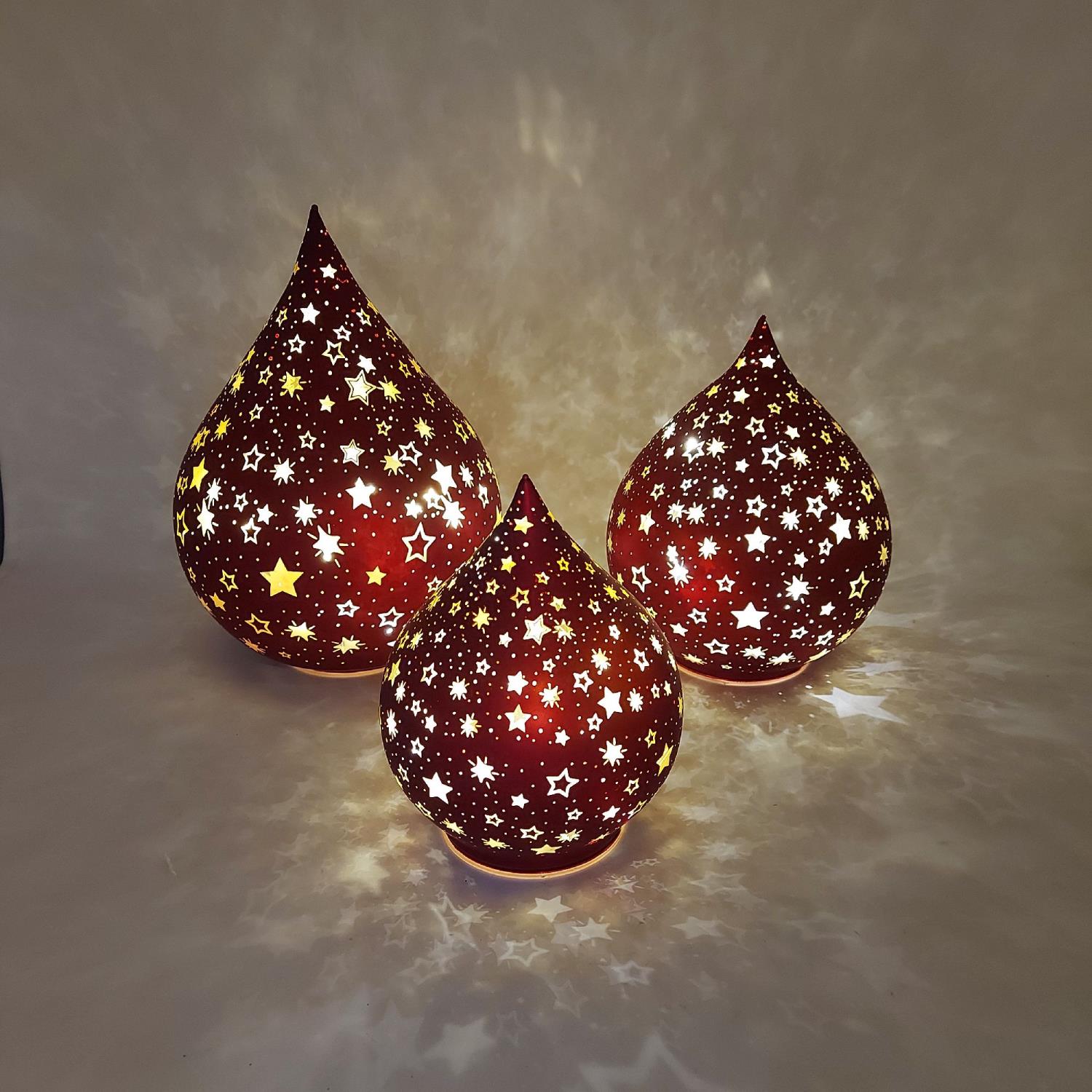 Drei rot leuchtende Teelichthalter in Tropfenform mit goldenen Sternen, die ein warmes Licht auf die