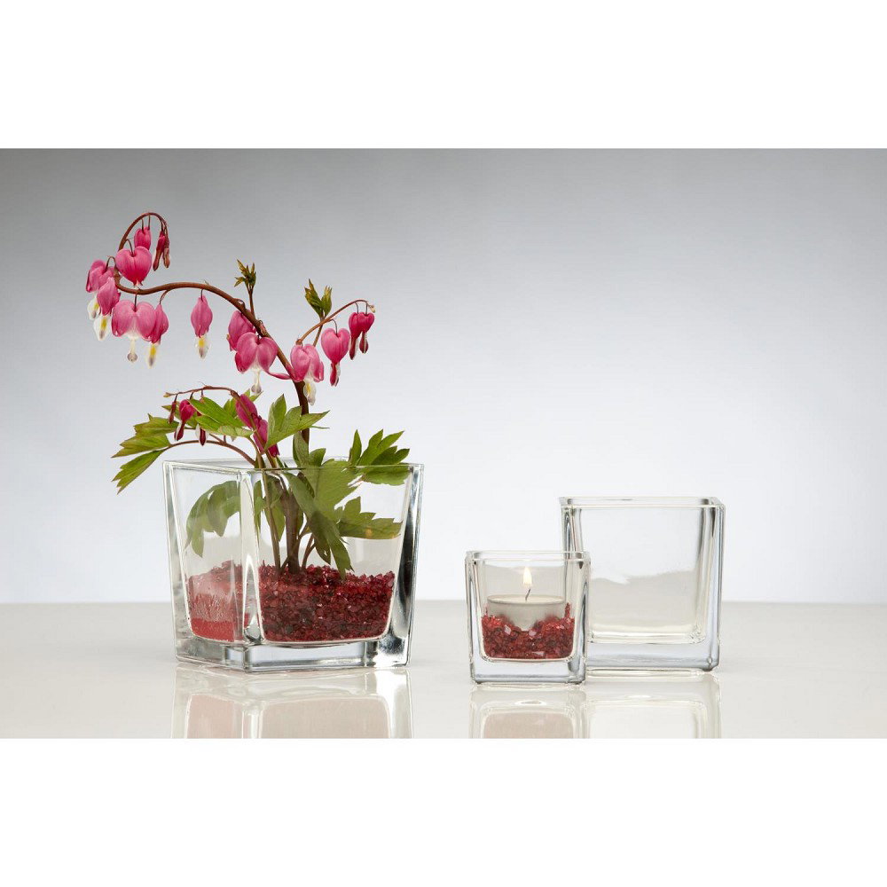 Ein Glas mit pinken Blüten und grünem Blattwerk steht neben zwei weiteren gläsernen Behältern, einer mit Kerze und einer leer.