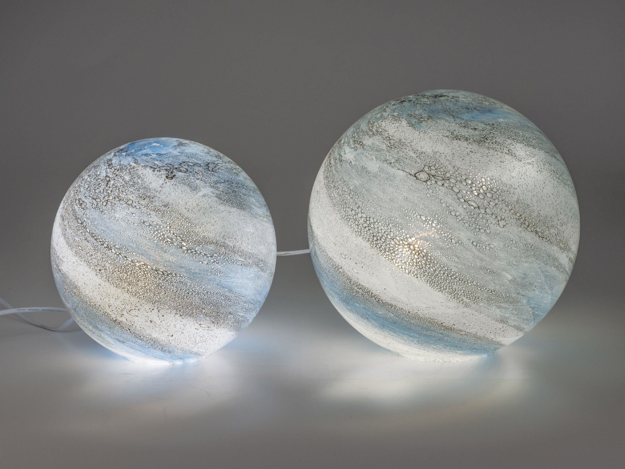 Zwei runde, gläserne Objekte in unterschiedlichen Größen, mit blauen und weißen Farbtönen sowie einer strukturierten Oberfläche.