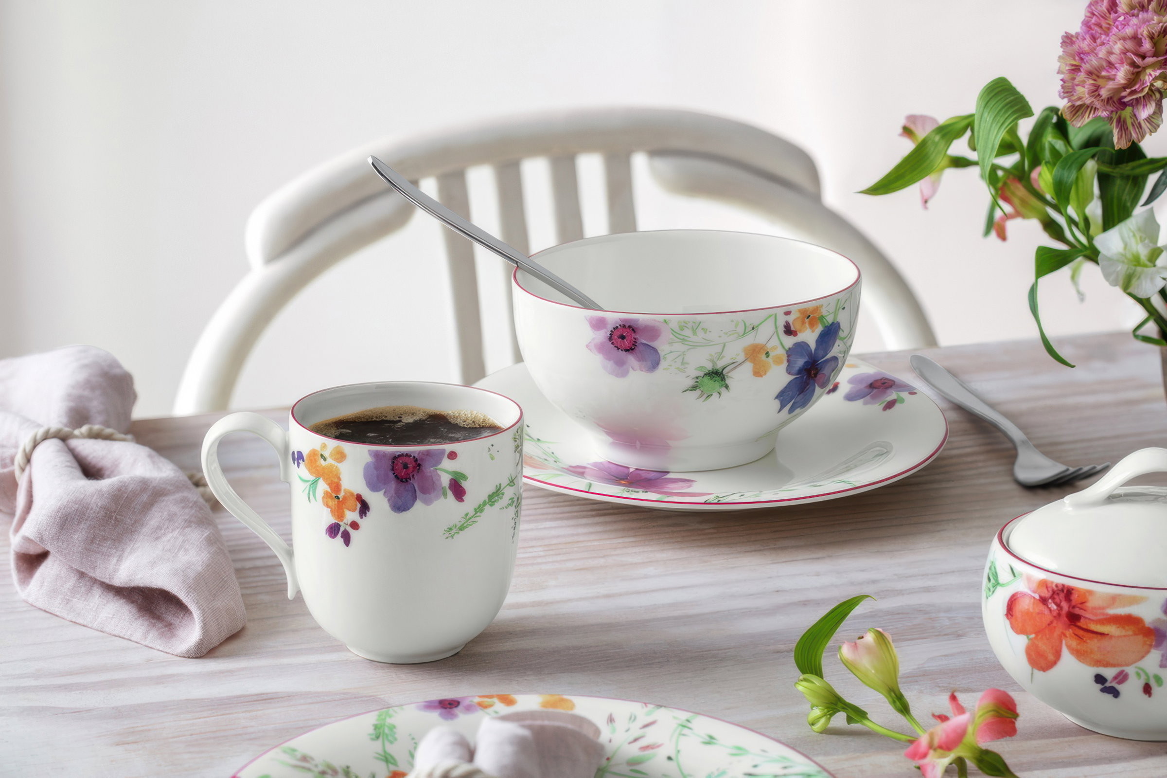 Tasse mit Kaffee und eine große Schüssel auf einem Tisch, umgeben von einem Löffel, einer Serviette und einer Blumenvase.