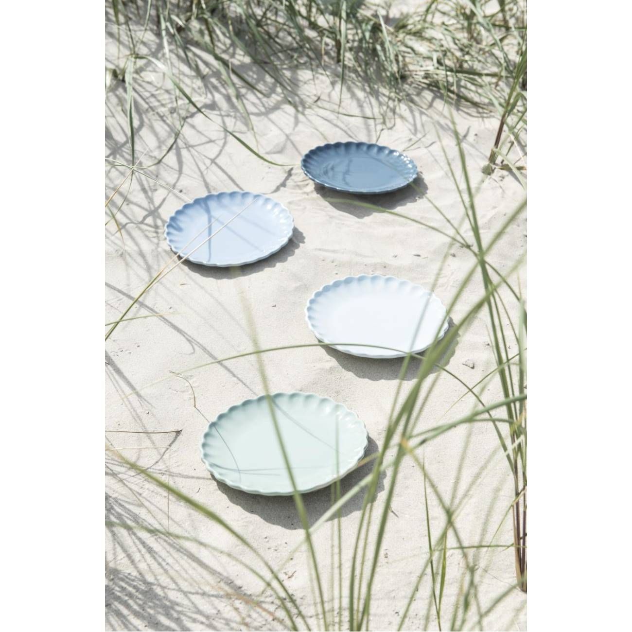 Fünf Teller in verschiedenen Blau- und Grüntönen liegen im Sand, umgeben von Gras.