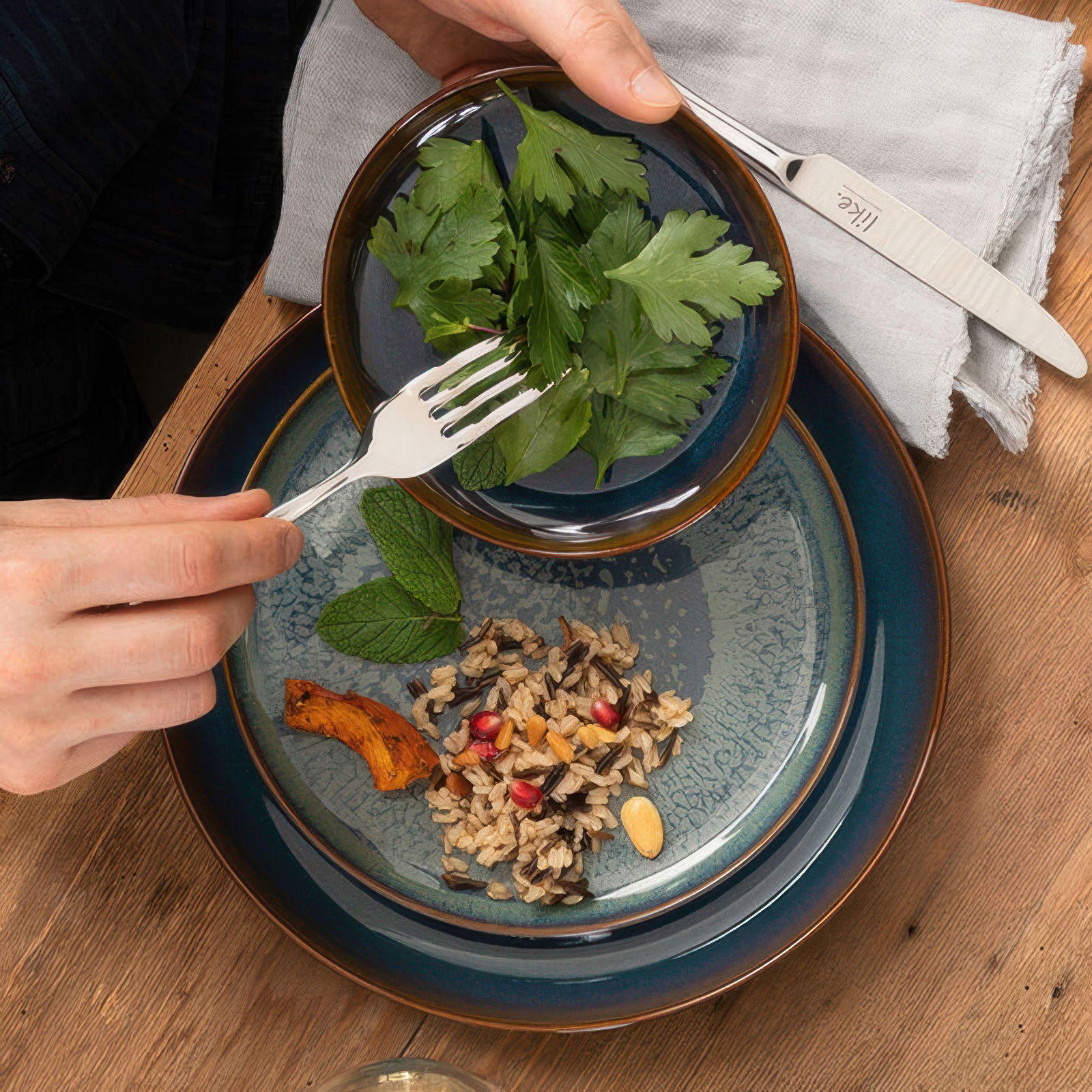 Eine Hand hält eine Gabel über einer Schüssel mit frischen Kräutern. Im Hintergrund sind verschiedene Teller mit Reis und Gemüse.